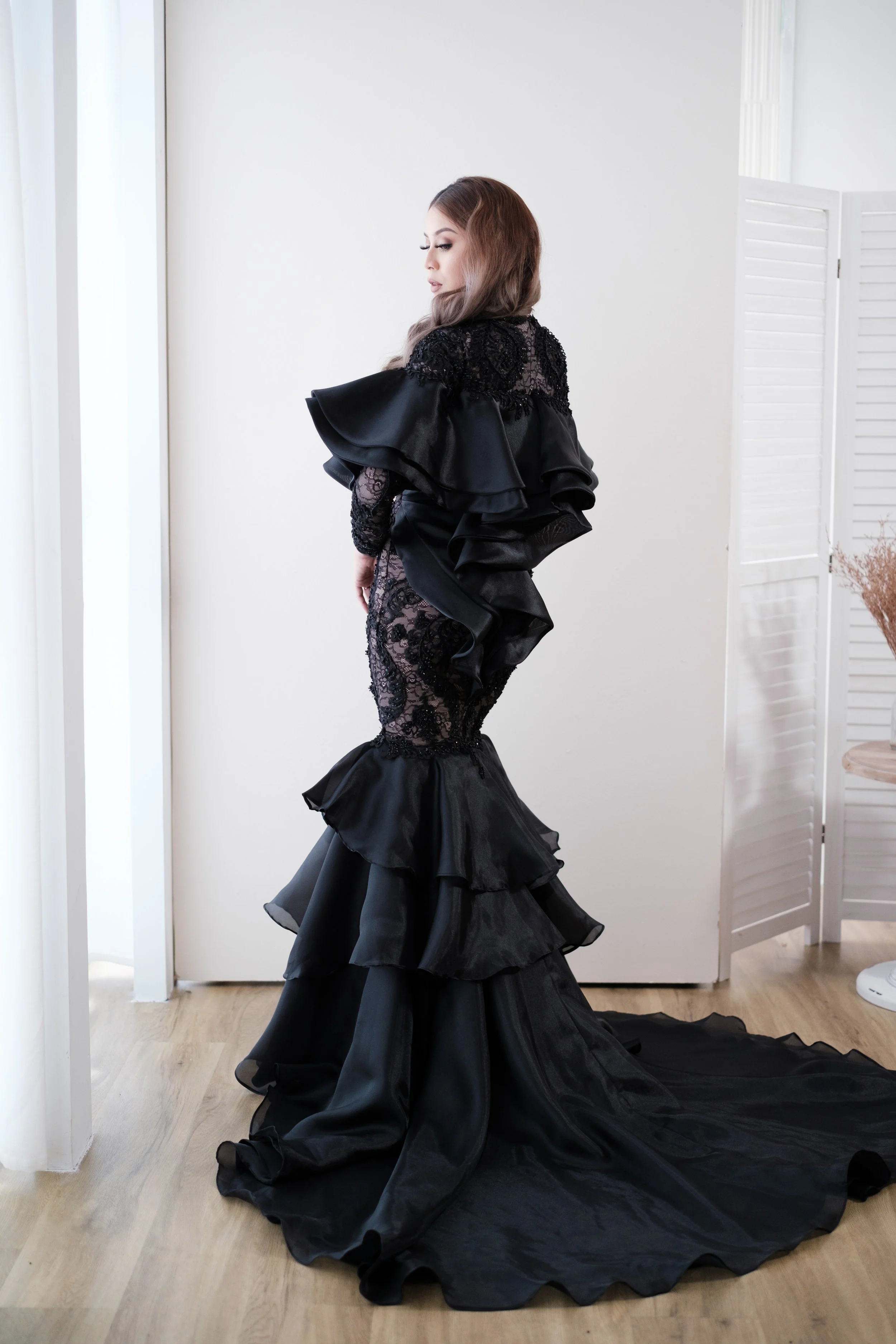 A woman with long brown hair is dressed in a black lace gown with ruffles and a long train, standing indoors on a wooden floor near a white wall and a white folding screen.