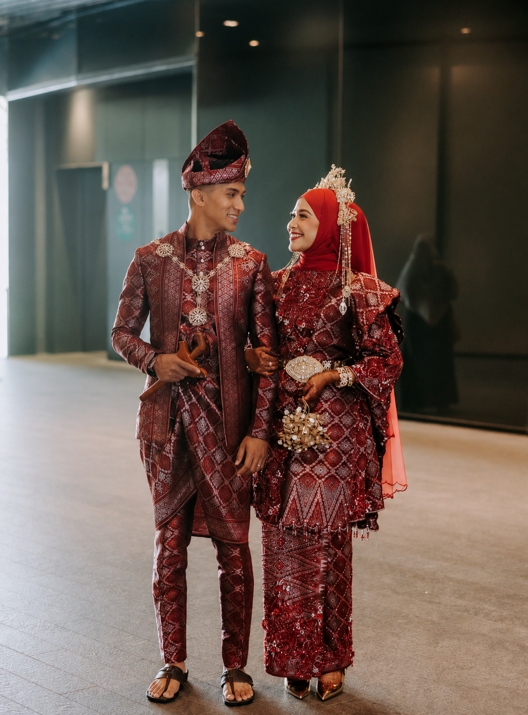 A newlywed Indonesian couple dressed in traditional red wedding attire, walking arm-in-arm and smiling at each other.