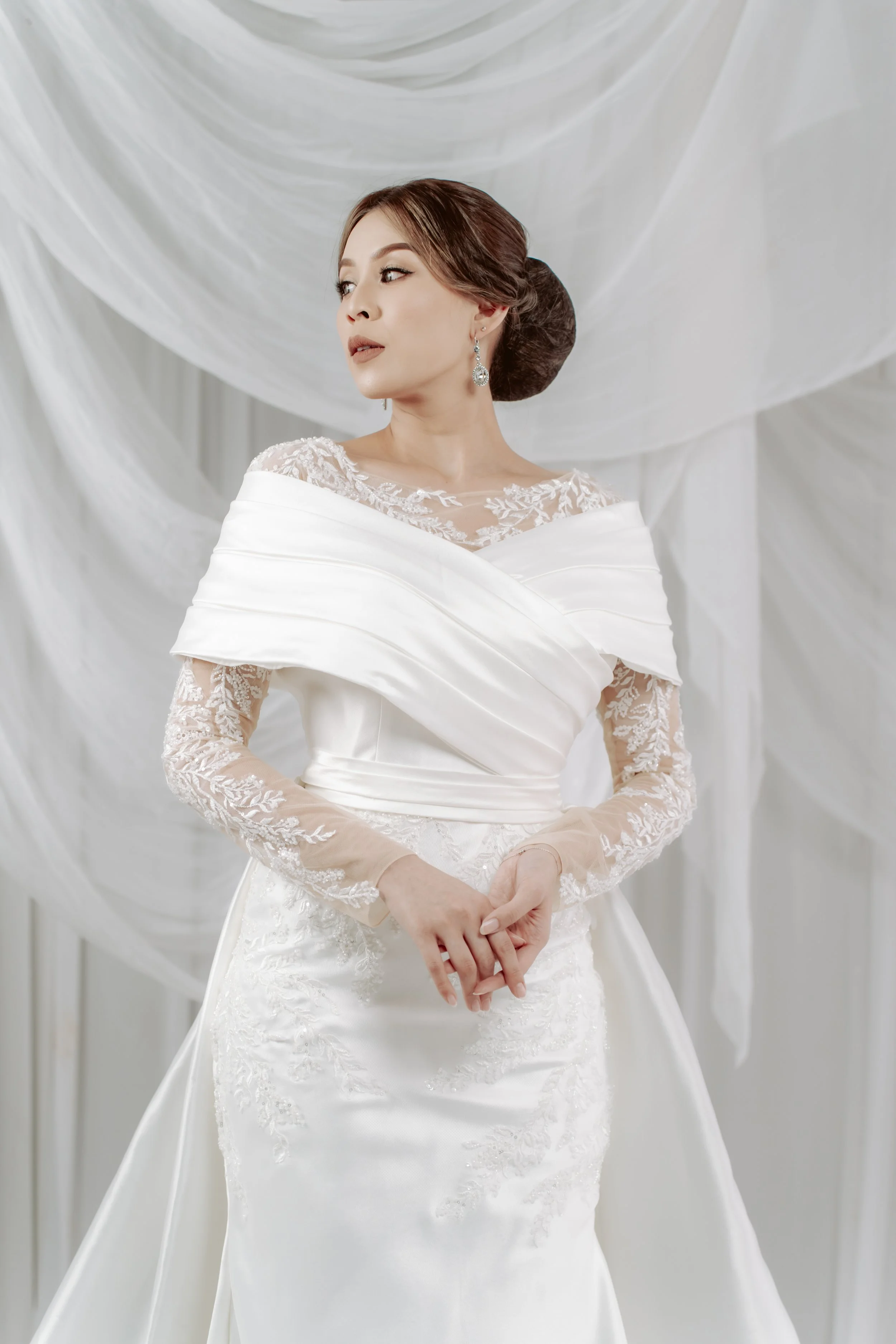 A woman in a white wedding gown with lace details and off-the-shoulder design, standing in front of draped white fabric.