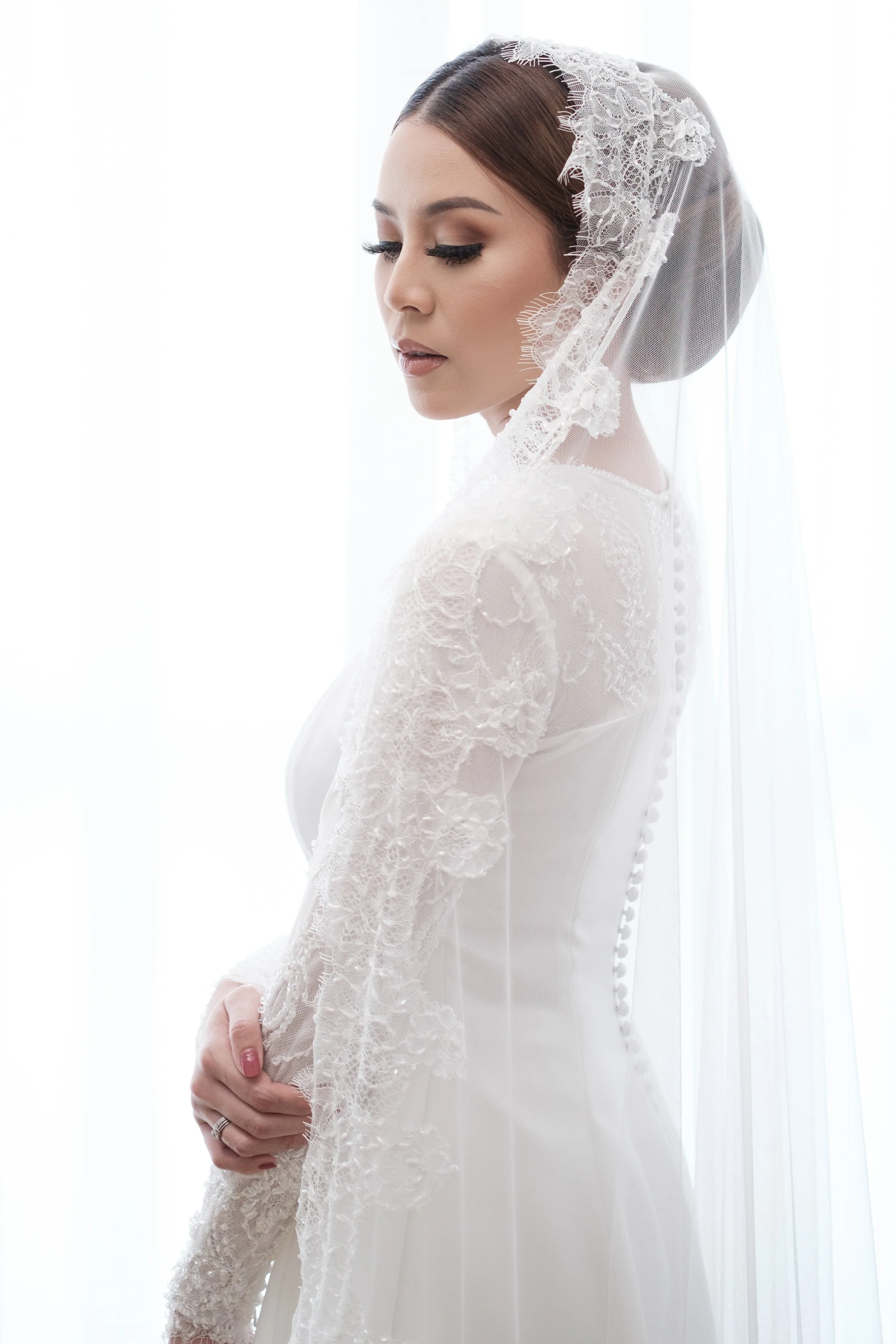 A bride with brown hair wearing a white lace wedding dress and a lace veil, standing in front of a bright white background with her eyes closed and hands clasped in front of her.