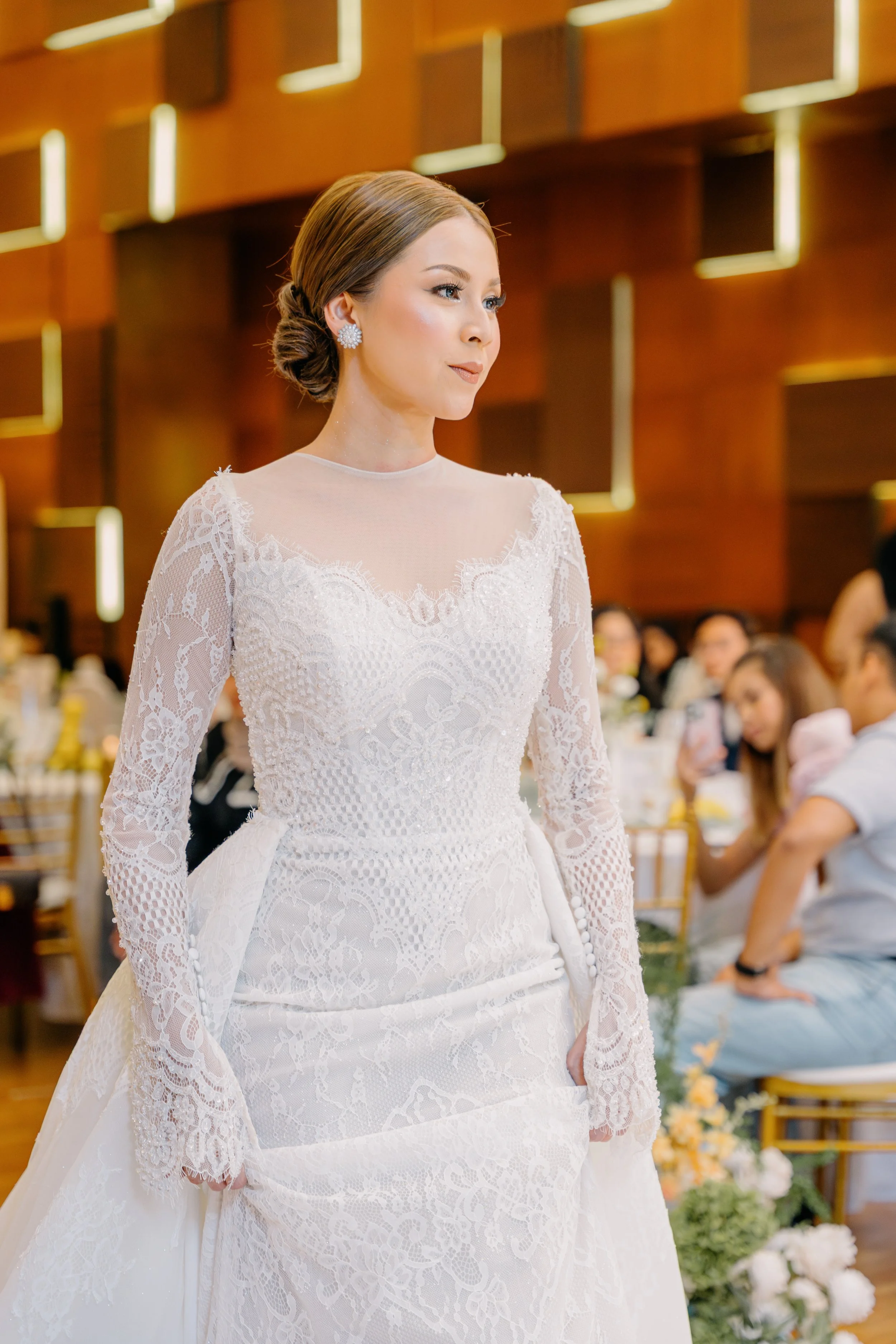 A woman dressed in a wedding gown with lace details, wearing large earrings and a bun hairstyle, at a wedding reception.