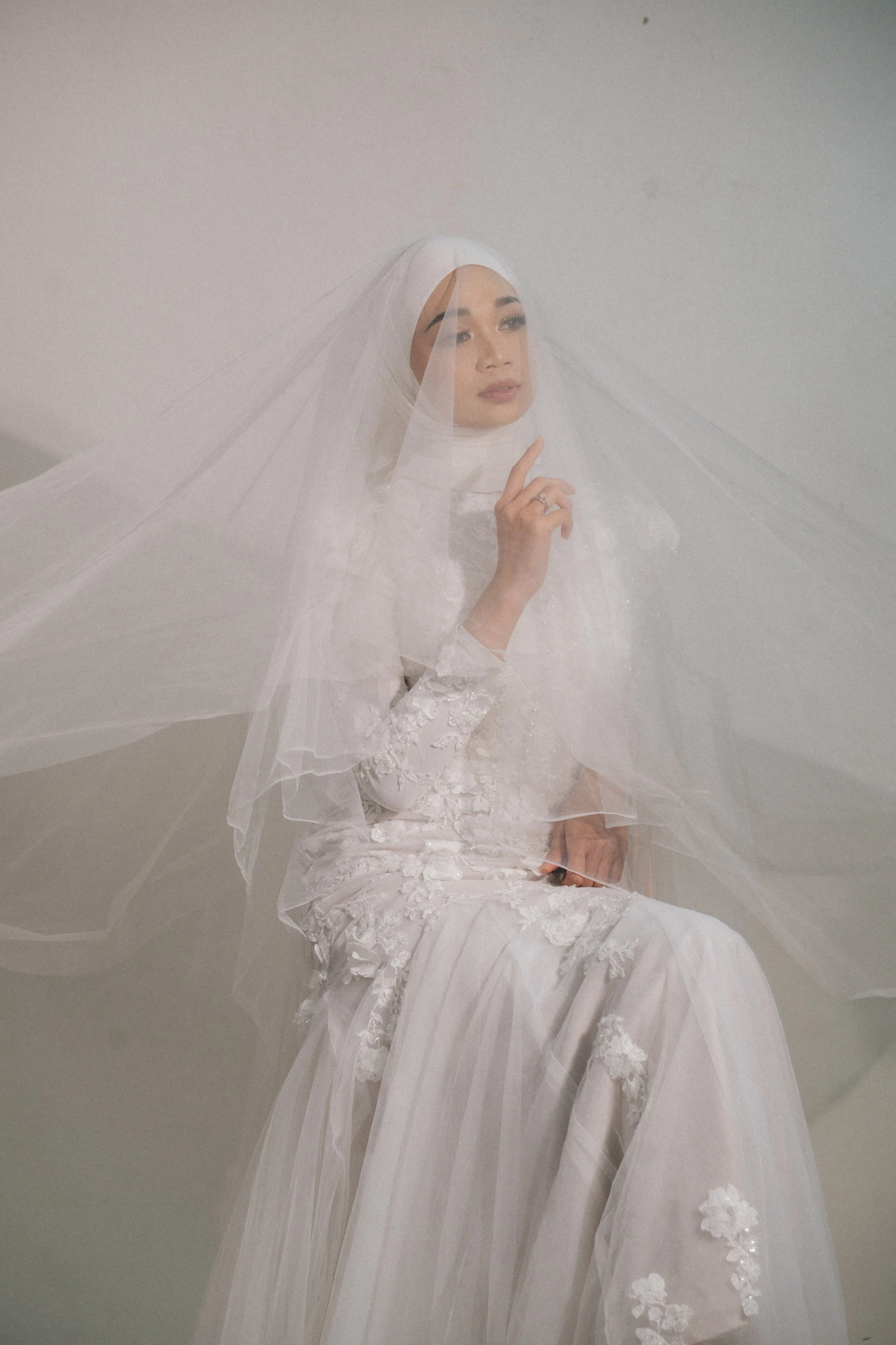 A woman in a white dress, veil, and hijab, sitting against a plain wall, with one hand near her face, looking thoughtfully to the side.
