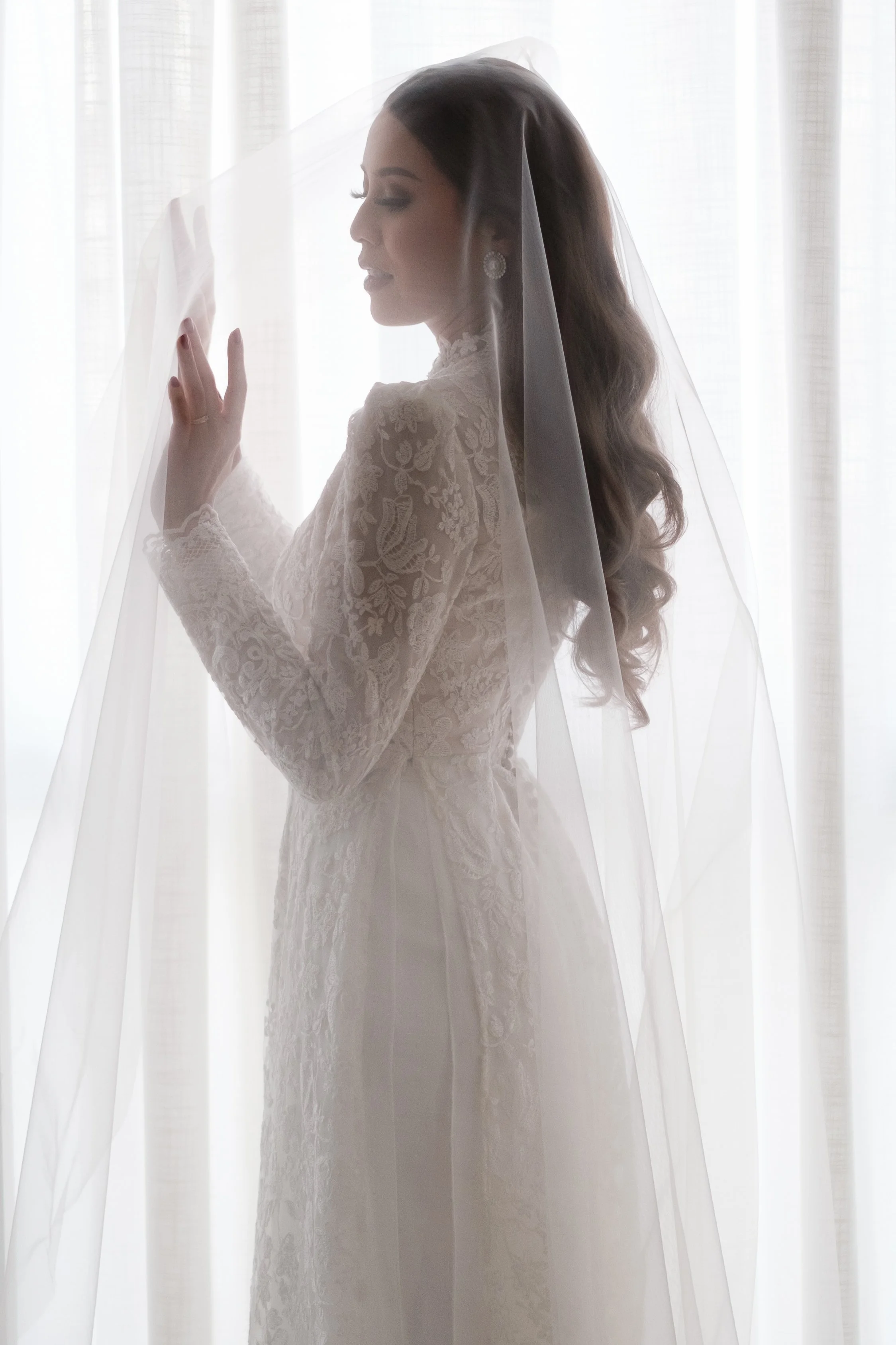 A bride in a lace wedding dress under a sheer veil, standing in front of backlit curtains. She is touching the veil with her left hand and has her eyes closed, smiling softly.