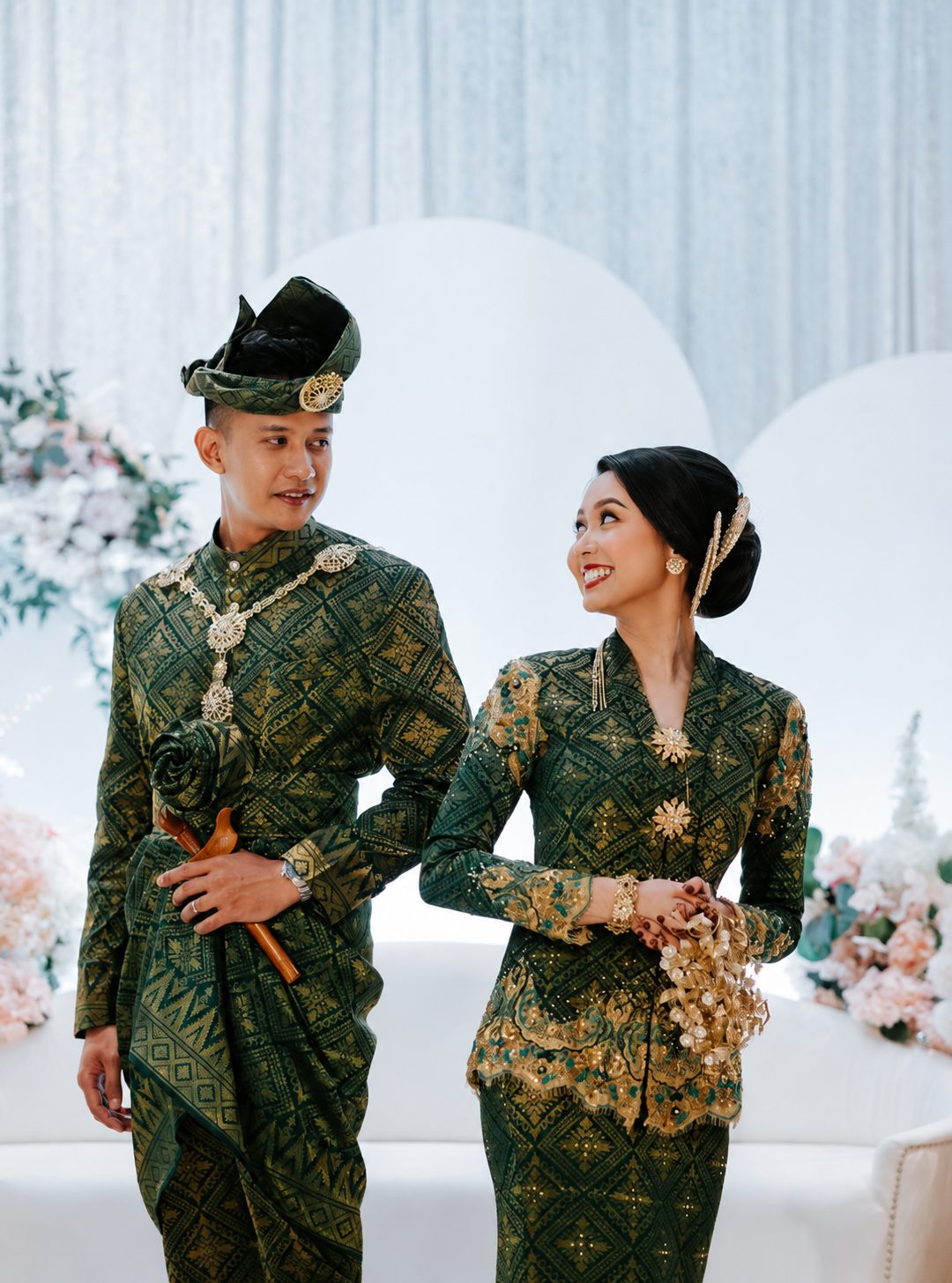 A man and woman dressed in traditional songket attire, standing indoors with floral decorations in the background, smiling at each other.