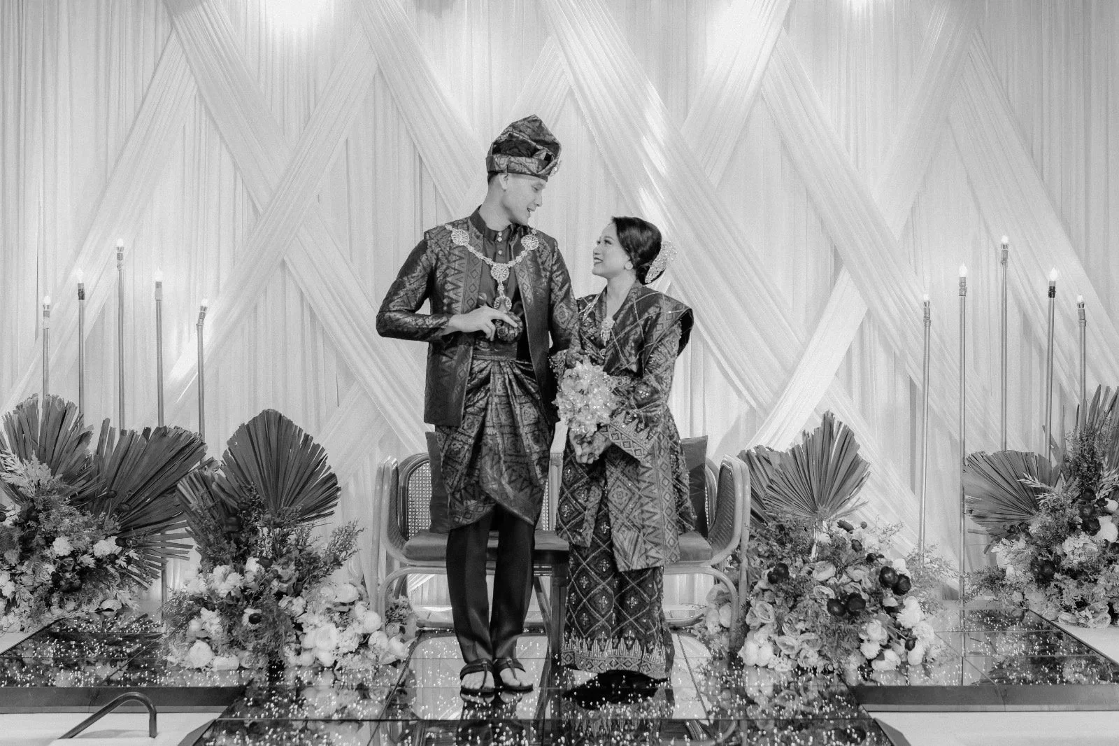 A black and white photo of a couple in traditional Indonesian wedding attire standing on a decorated stage, surrounded by floral arrangements.