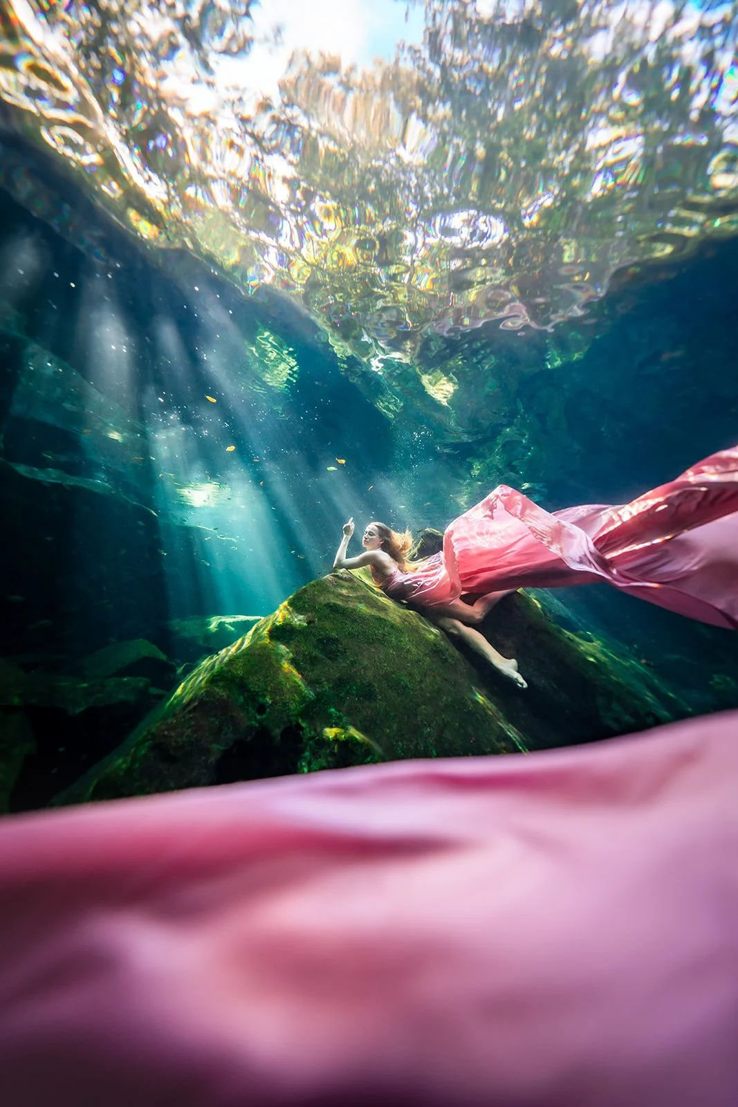 A white woman posing for an underwater photoshoot in a pink flying dress in a Mexican cenote