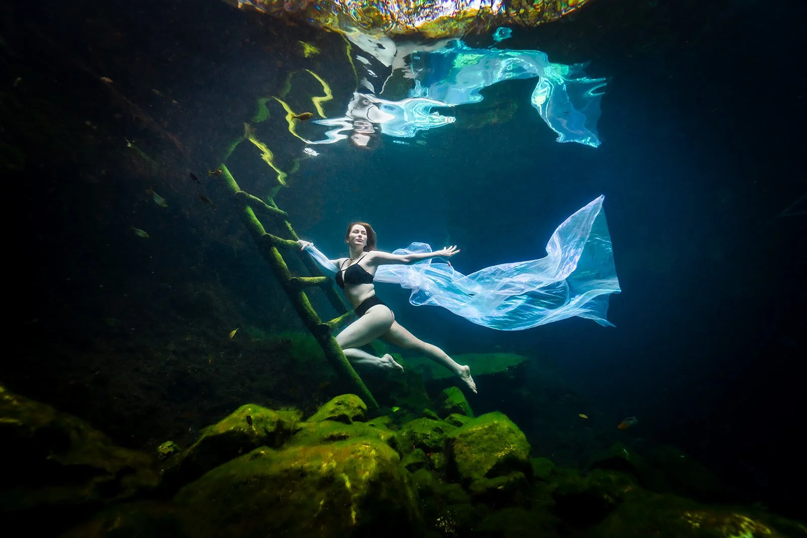 A white woman posing next to a later for an underwater photoshoot, wearing a black bikini