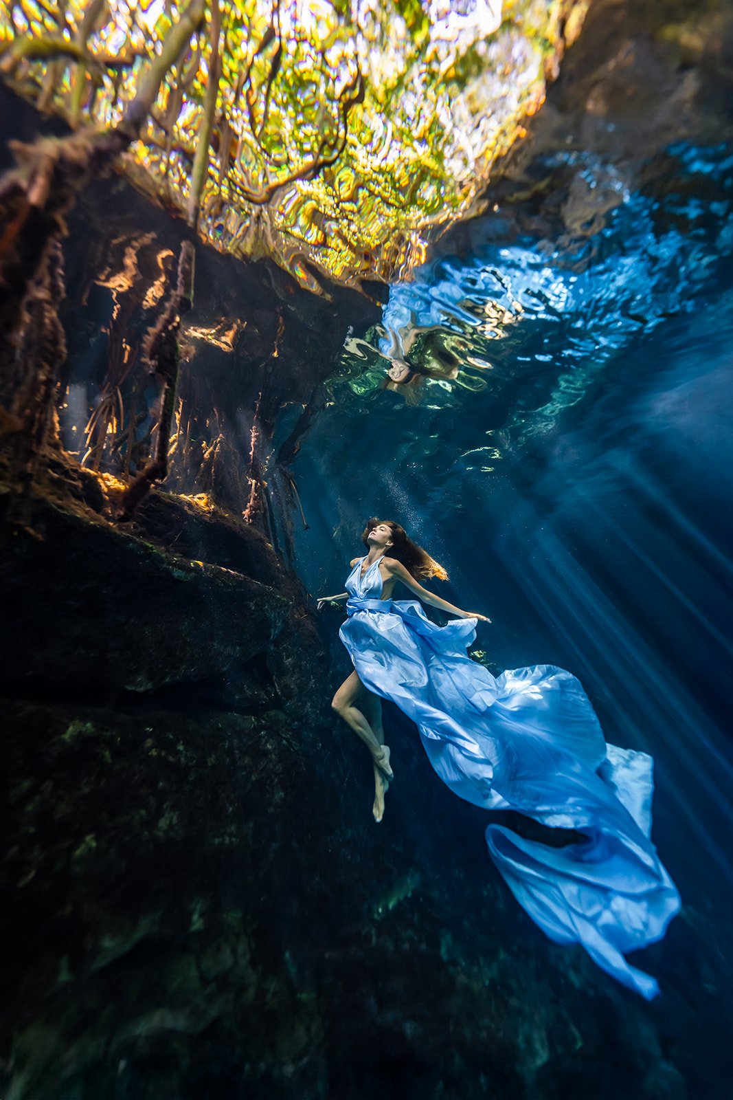 A woman modeling for an underwater photoshoot wearing a blue flying dress in a cenote in Tulum