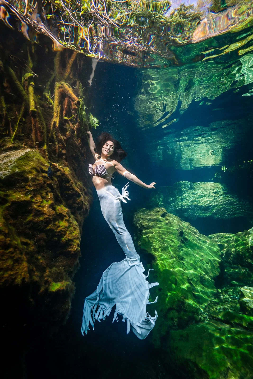 An Asian woman posing for an underwater photoshoot wearing a white mermaid tail in a crystal clear cenote in playa del Carmen
