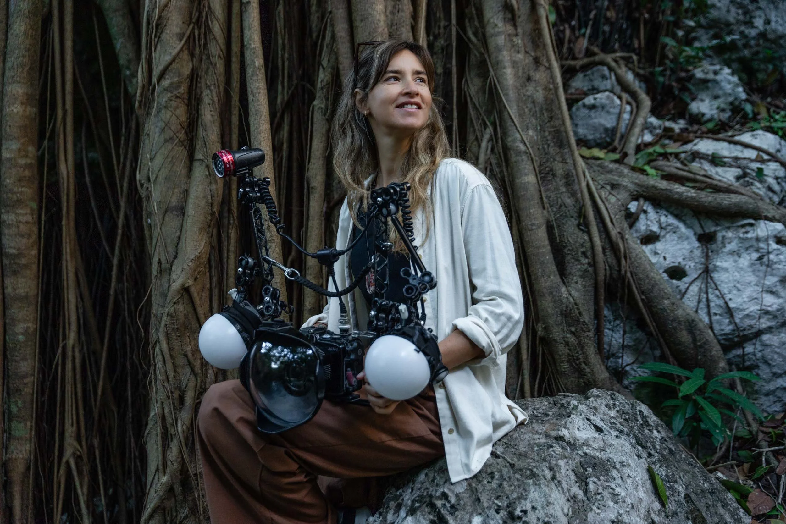Person holding an underwater camera setup while sitting on a rock in a forest with tree roots in the background.