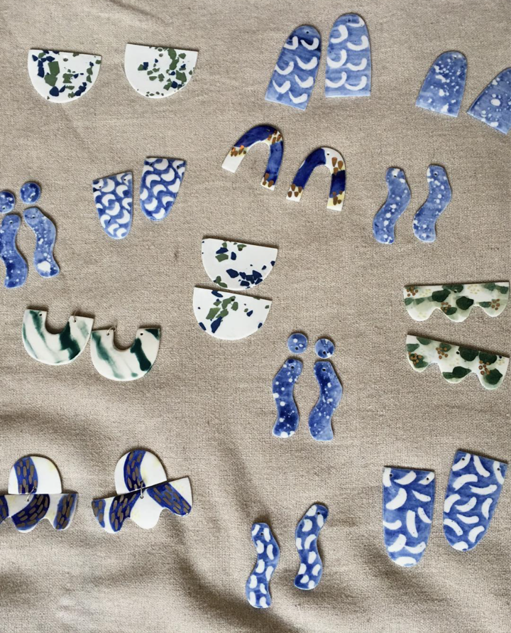 Various small, decorated ceramic or plaster pieces arranged on a beige fabric surface. The pieces are semicircular or curved, decorated with blue, green, and white patterns, resembling abstract or stylized designs.