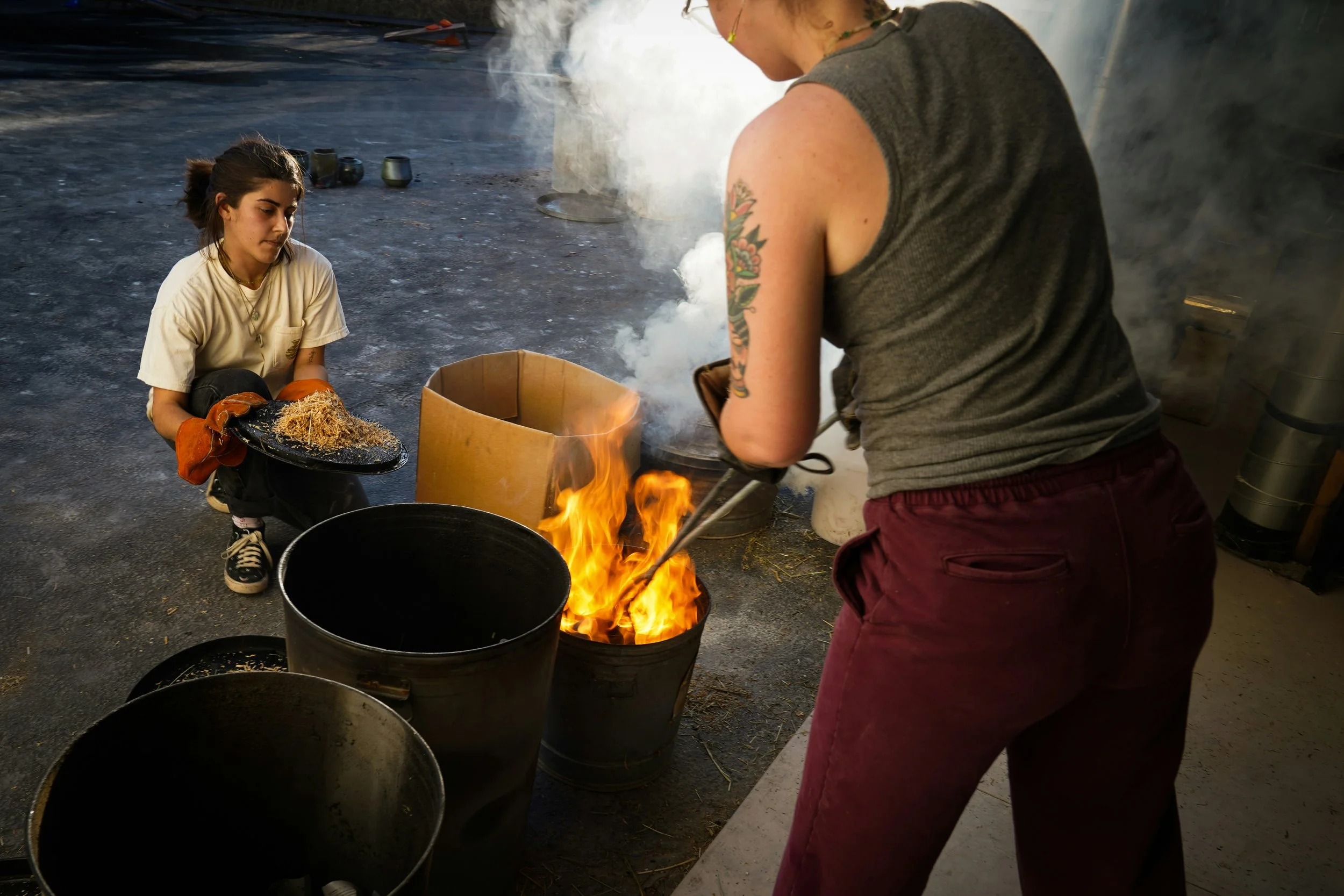 Two women working with fire and black buckets. One is crouching with a tray of wood shavings, wearing gloves, and the other is standing, handling a metal tool near the flames.