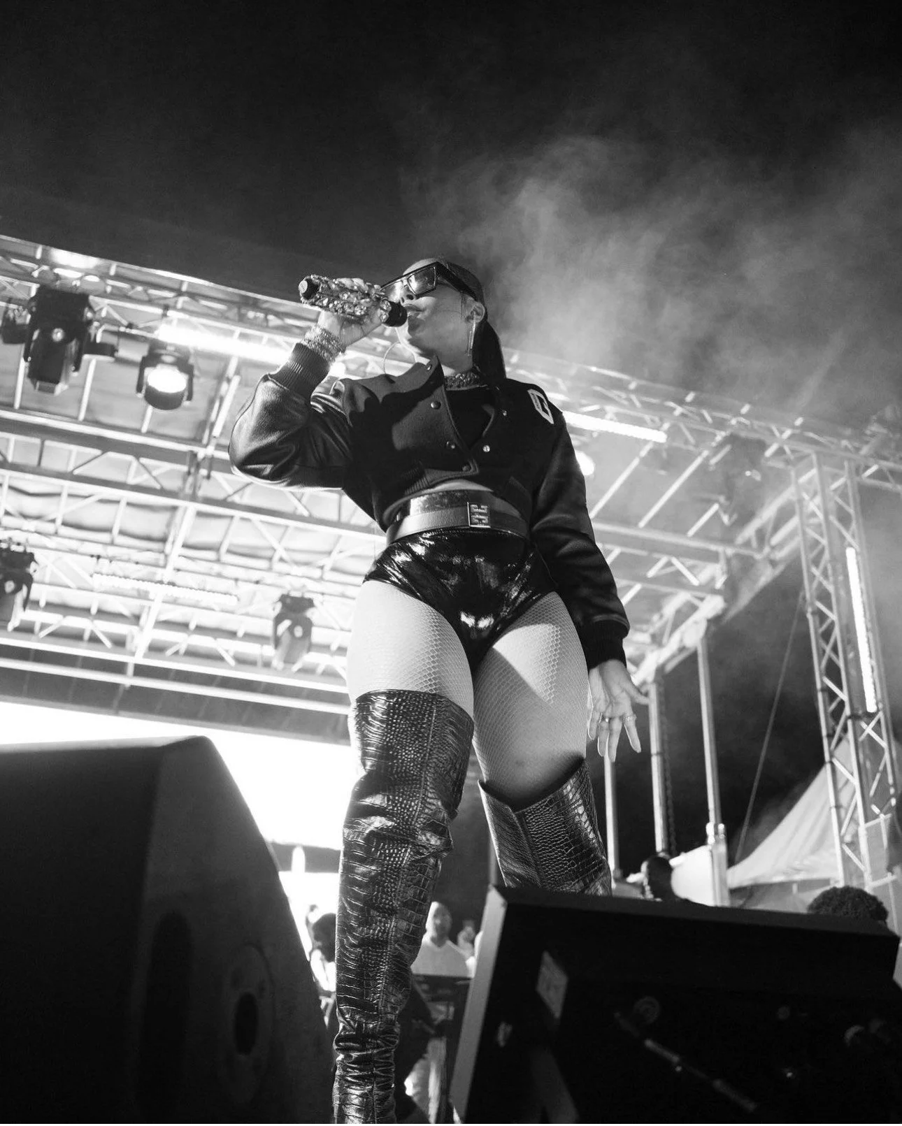 A female performer on stage drinking from a bottle, wearing a black jacket, shiny shorts, fishnet stockings, and thigh-high boots, with stage lighting and rigging in the background.