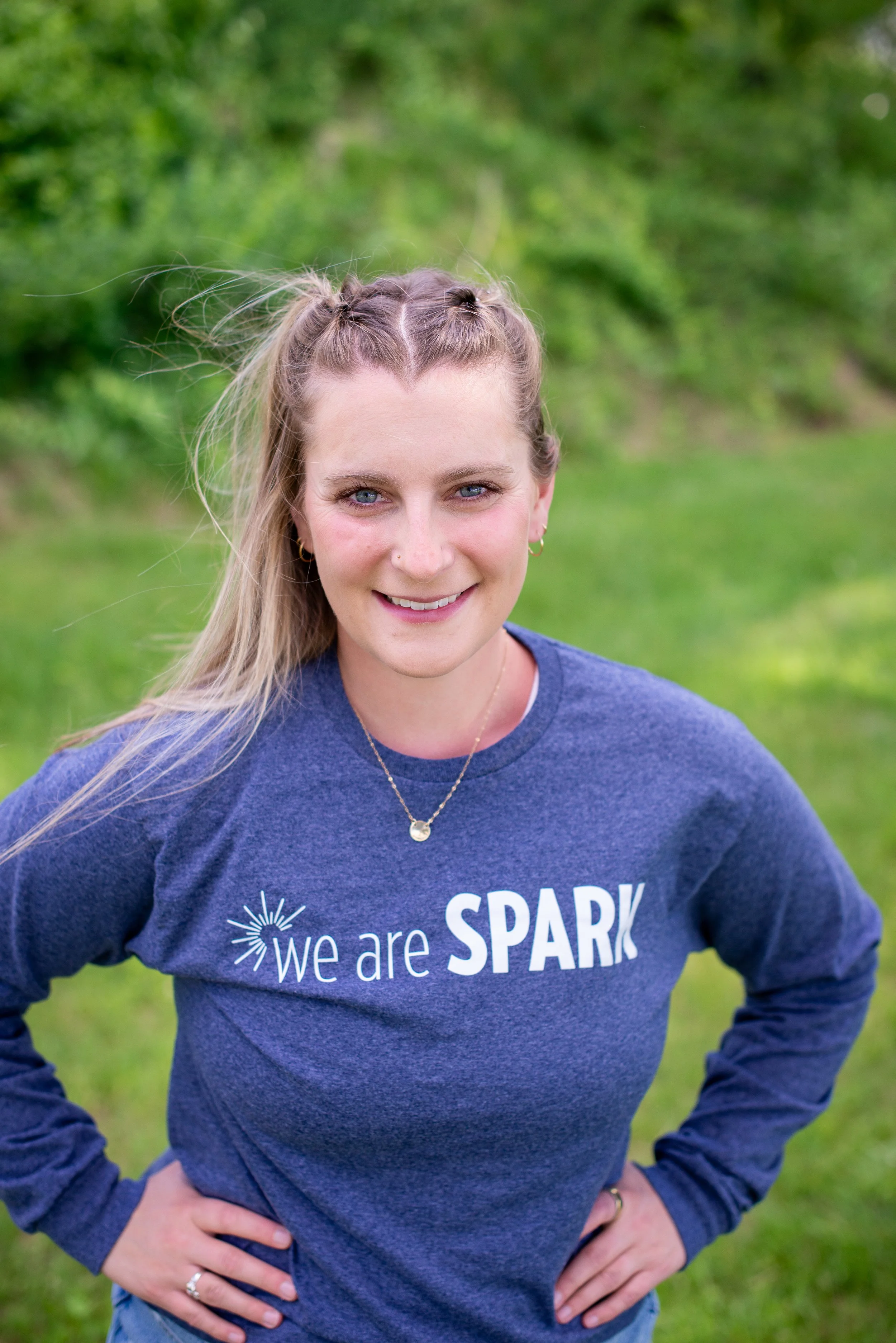 A smiling woman with blonde hair in a bun, wearing a black sleeveless top and a microphone headset, standing in front of a gym logo that reads 'SPARC CYCLING' on a light-colored wall.