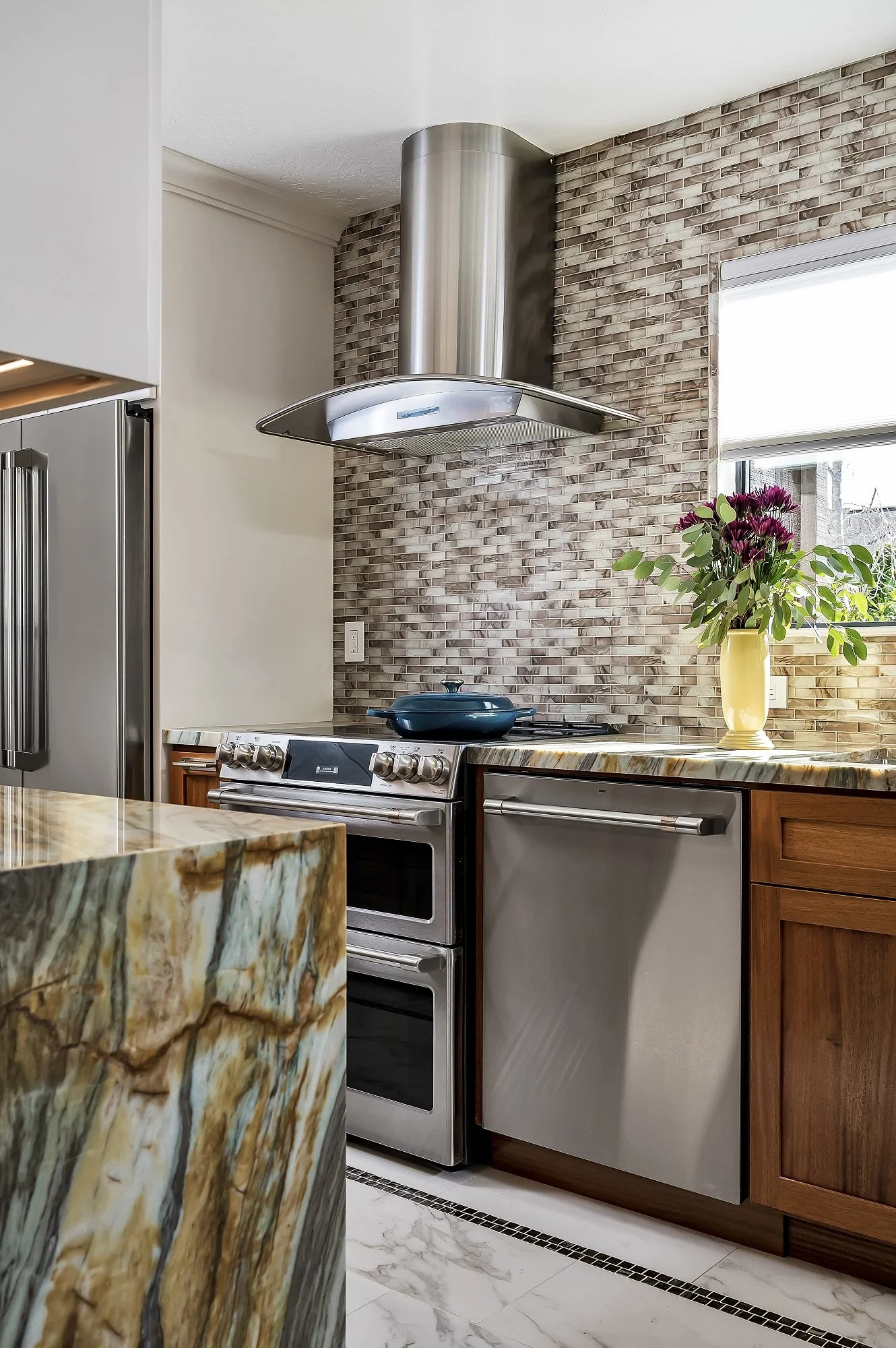 Custom condo kitchen remodel in Santa Cruz featuring wood cabinetry, a dramatic stone waterfall island, mosaic tile backsplash, and a modern stainless steel range with statement hood.