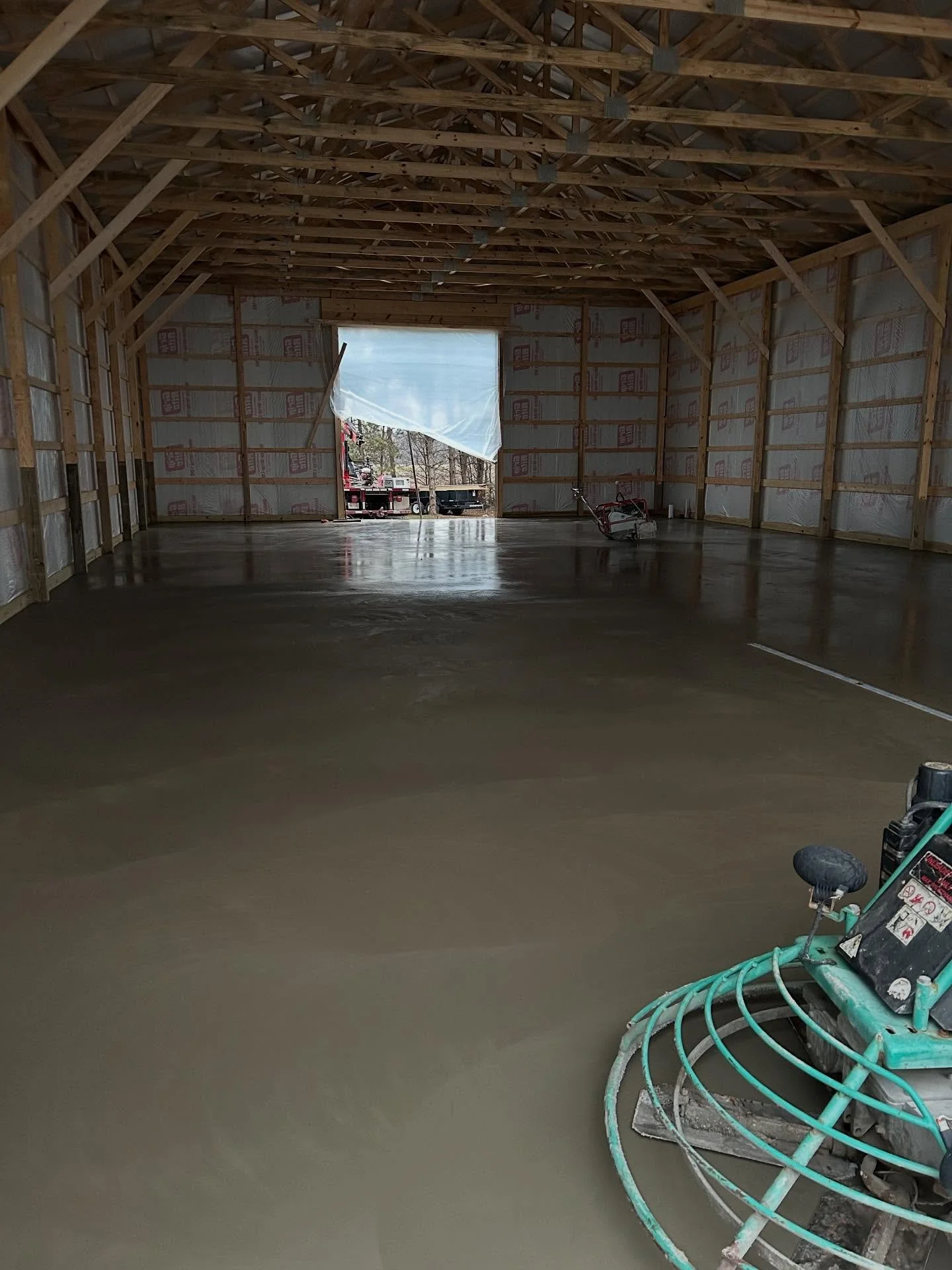 Interior of a barn under construction with a concrete floor being smoothed, exposed wooden framework, and an open garage door showing construction equipment outside.