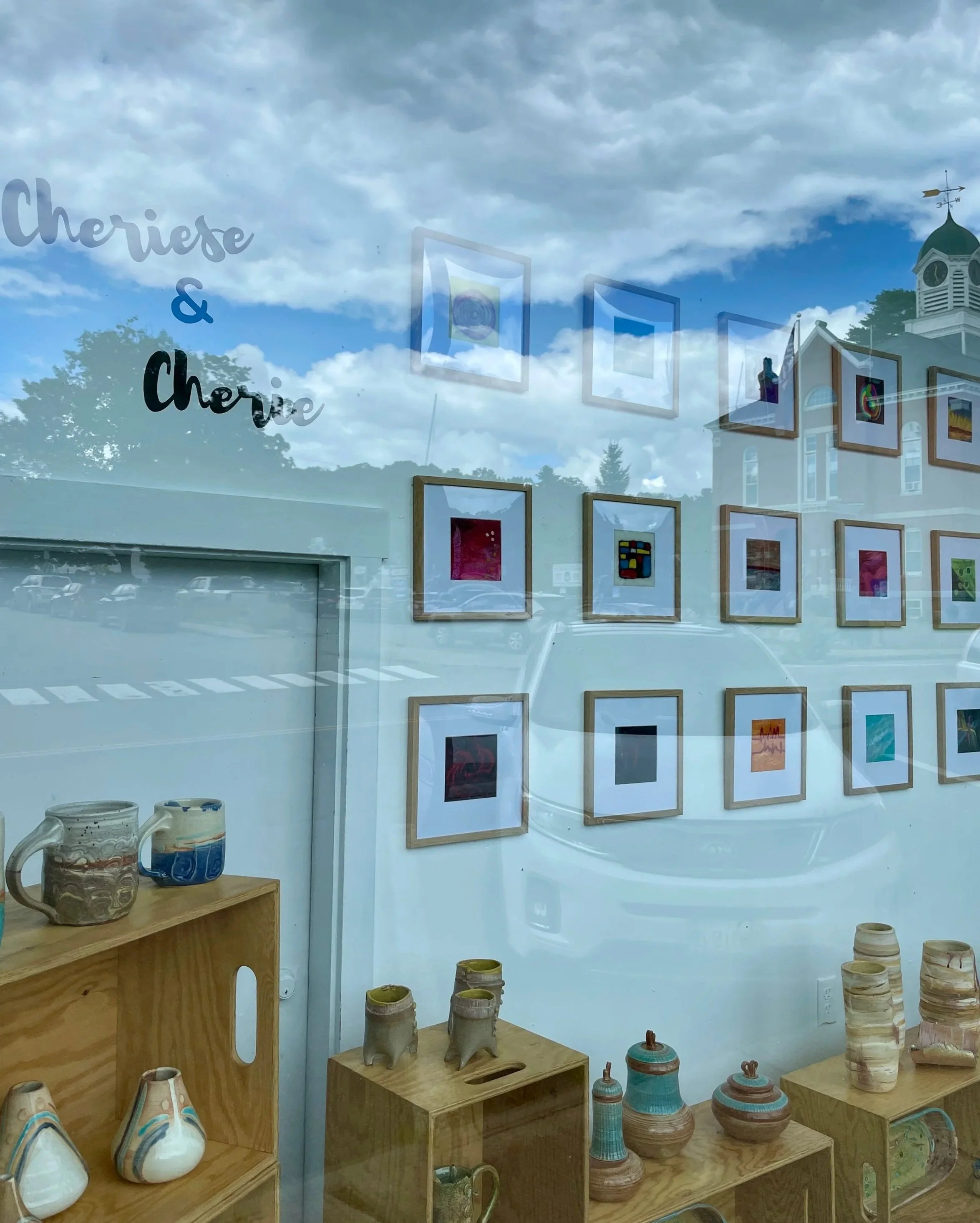 Art gallery window reflecting cloudy sky and trees, with framed artworks on the wall inside and pottery on wooden shelves.