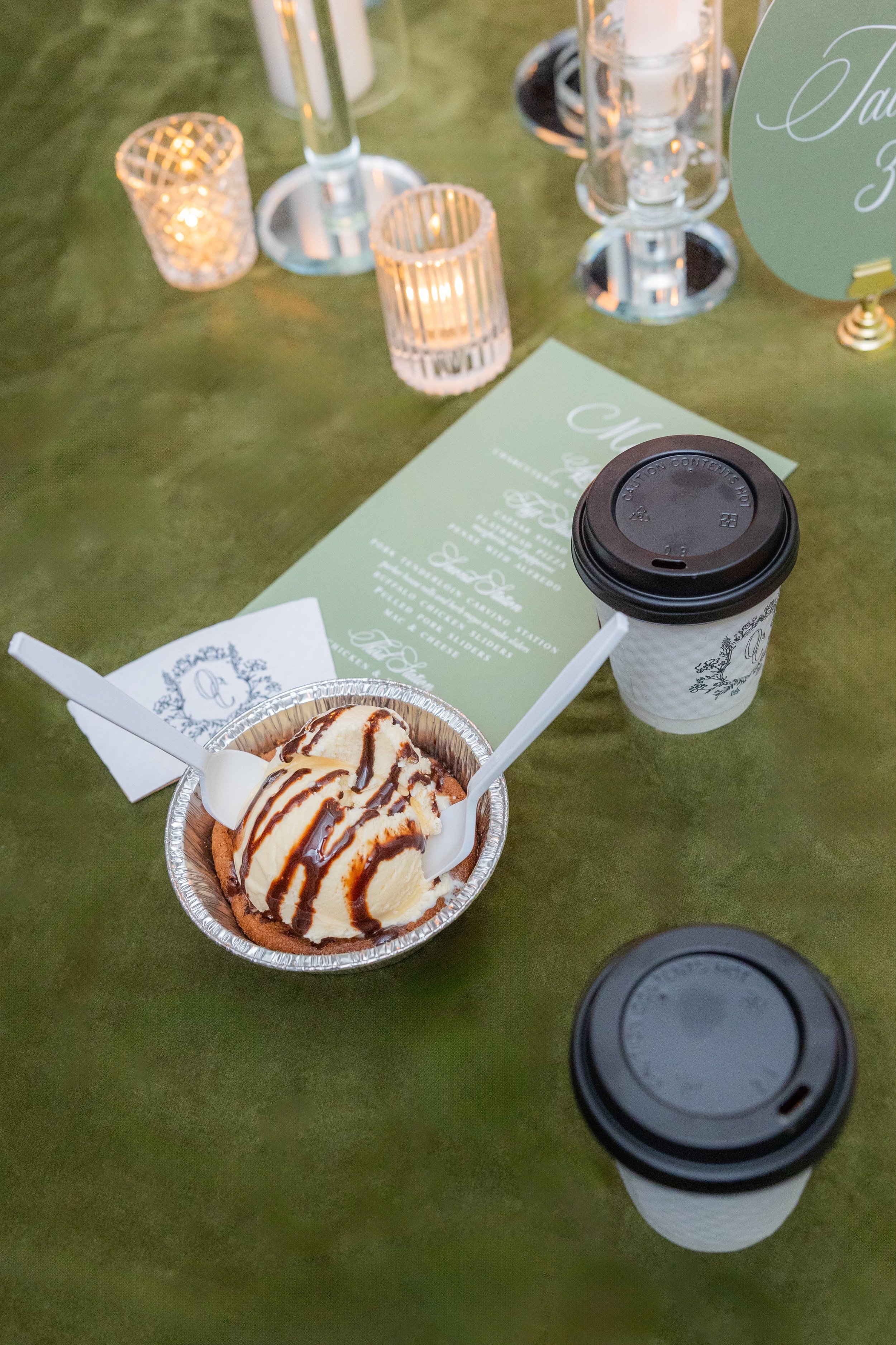 A dessert cup with vanilla ice cream drizzled with chocolate syrup and two white spoons, near two coffee cups with lids, on a green table with candles and a menu.