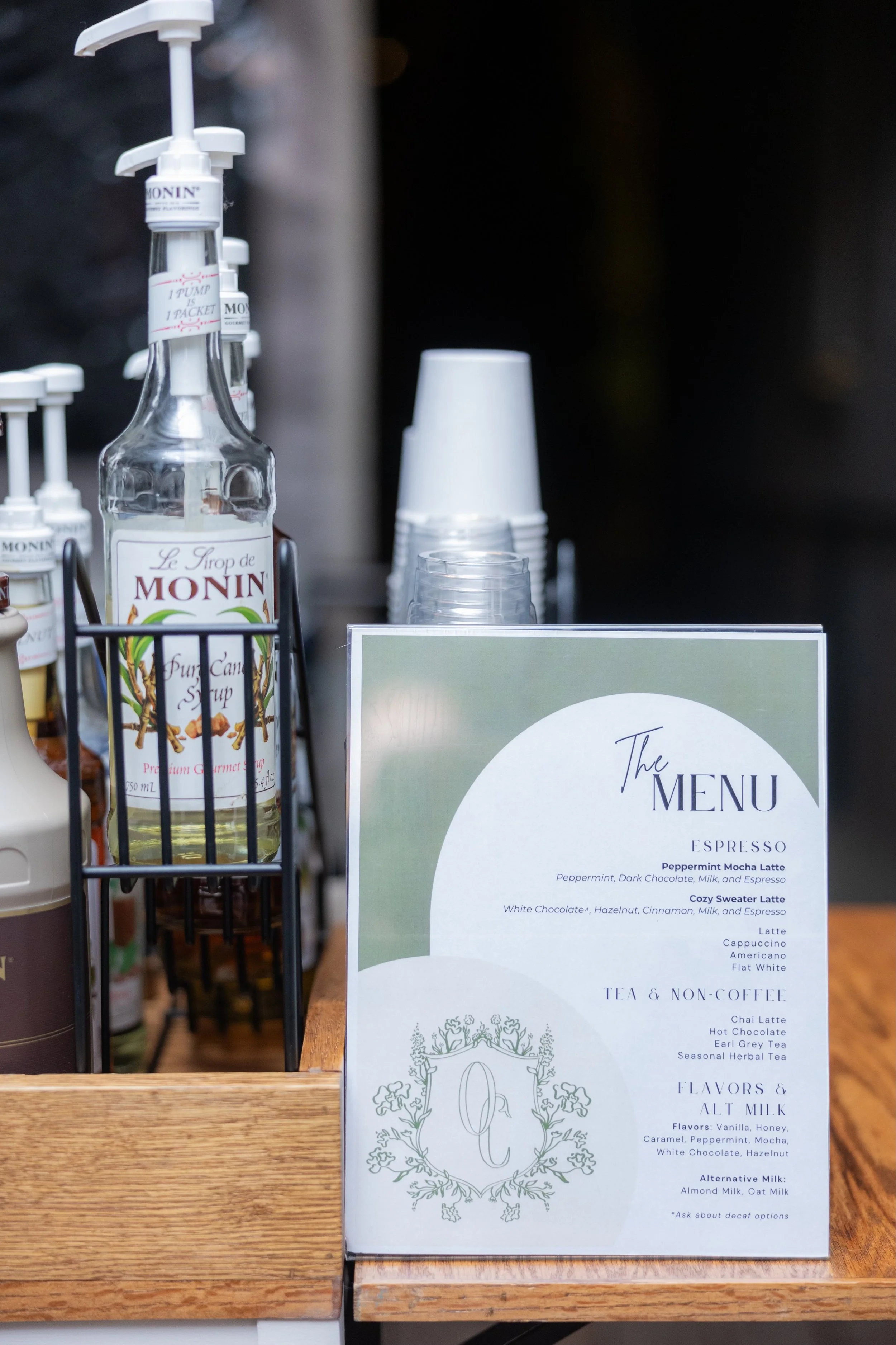 Coffee menu with espresso, tea, flavored, and alternative milk options, placed on a wooden counter next to syrup bottles and plastic cups.