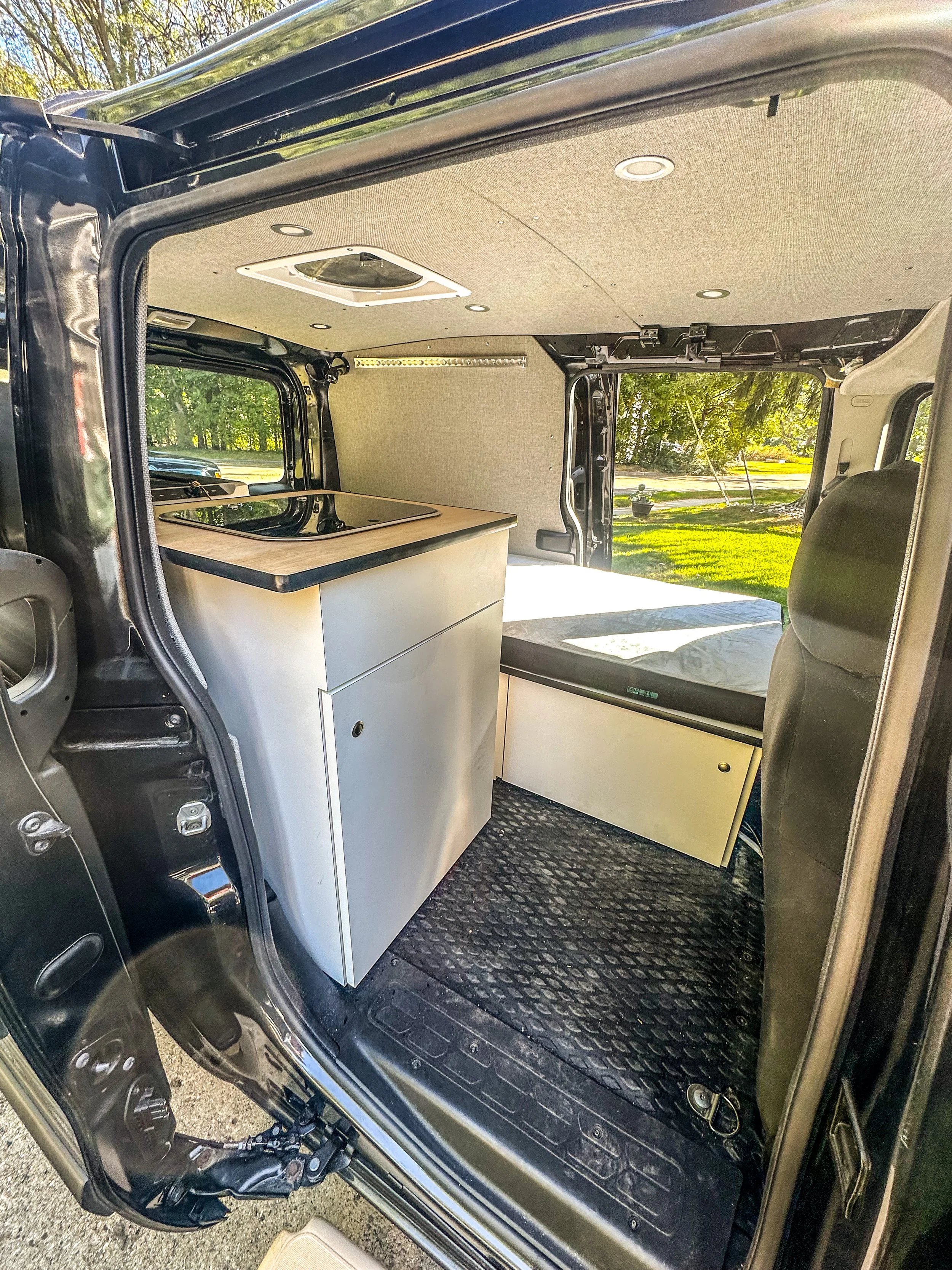 Interior of a converted van with a compact kitchen, including a stovetop and storage cabinets, with a bed at the back, in a grassy outdoor setting.