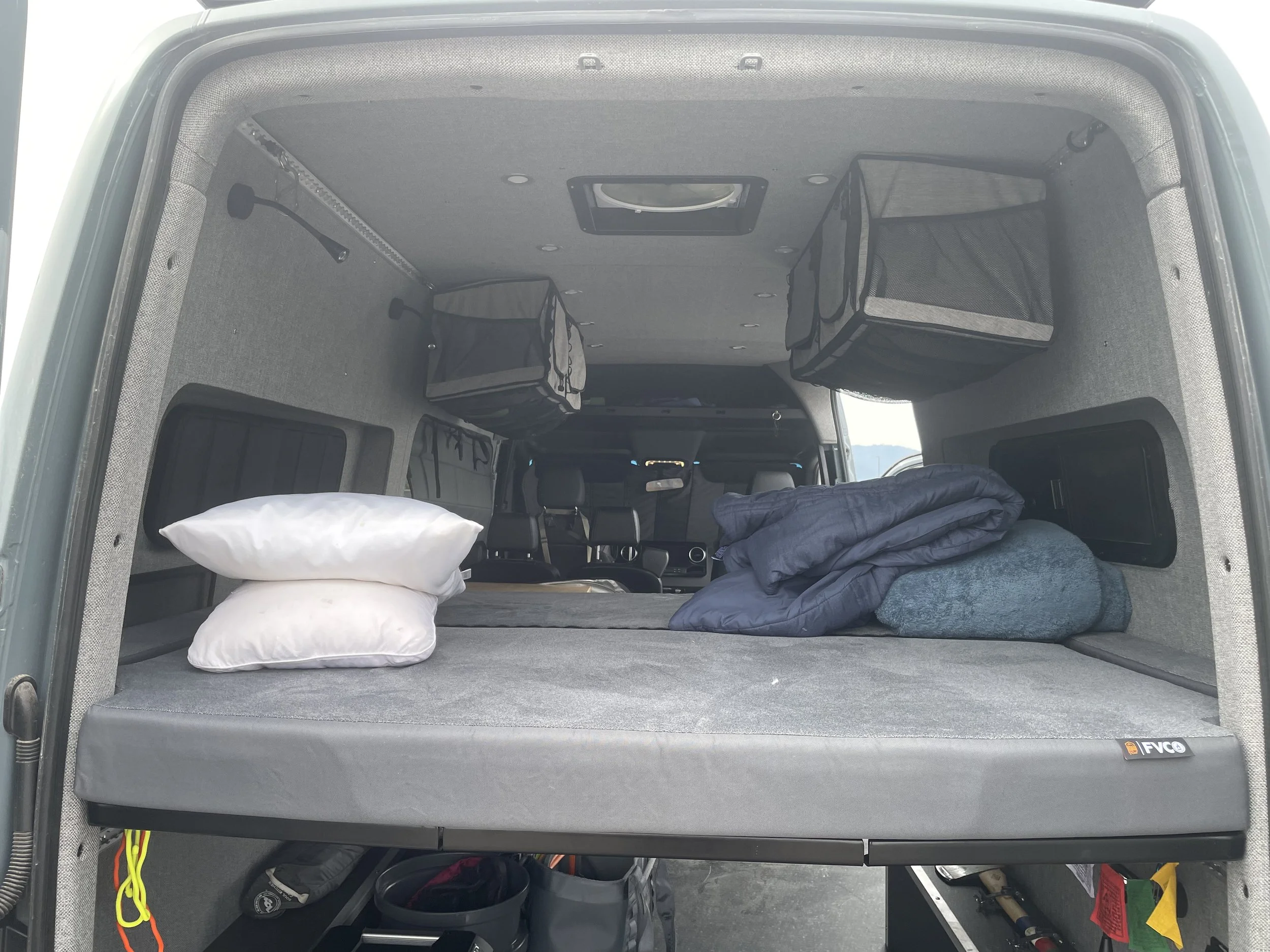 Interior of a van converted into a sleeping area with pillows, a rolled-up blanket, and storage containers.