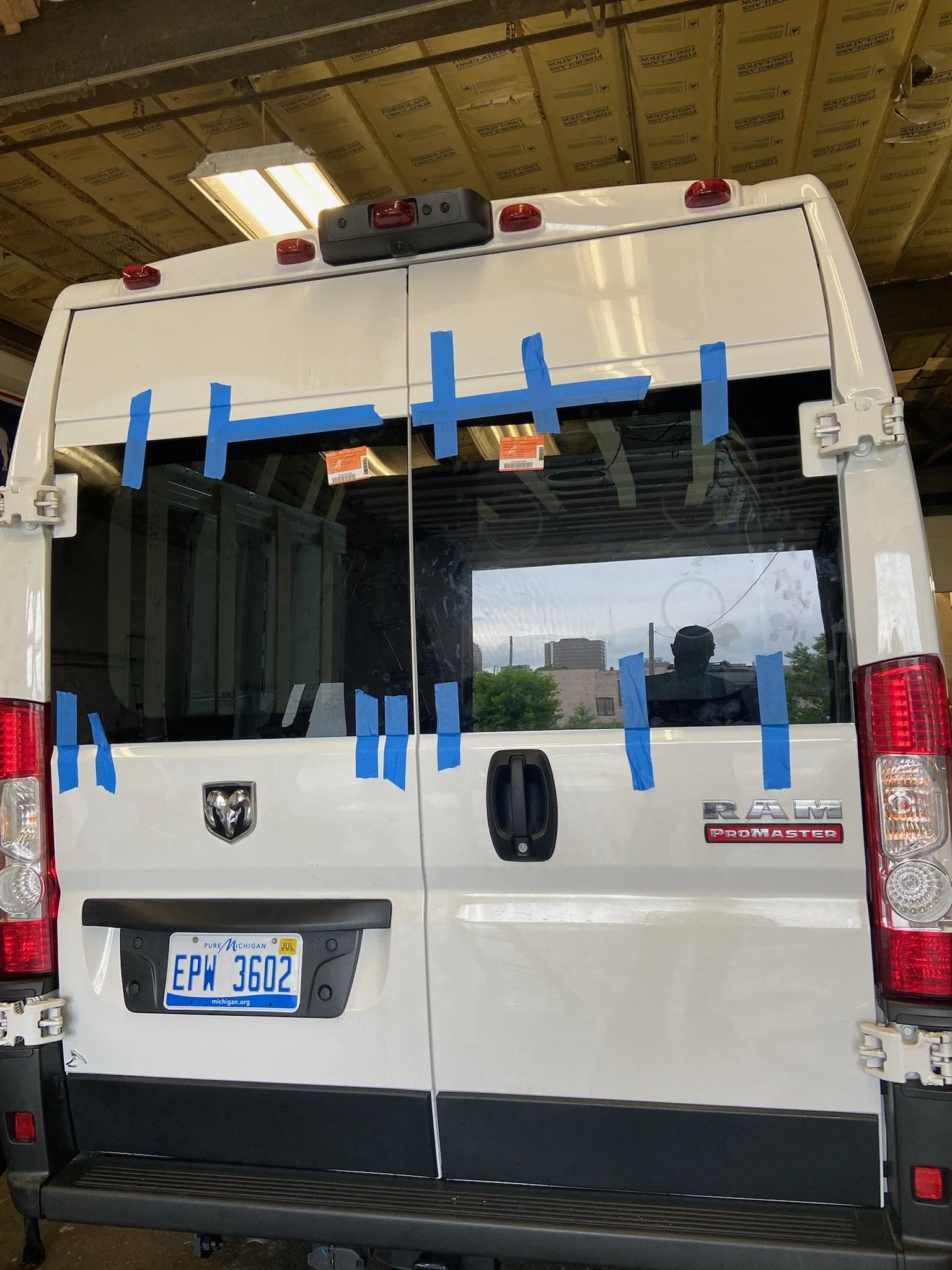 The back of a white Ram ProMaster cargo van with blue painter's tape applied in vertical and horizontal strips on the rear window and doors.
