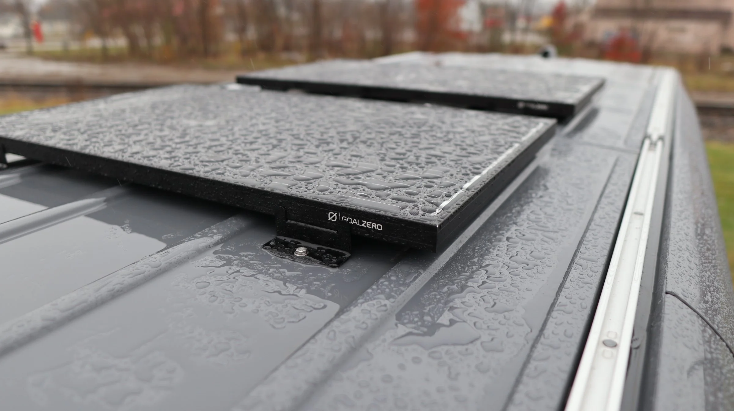 Close-up of a black solar panel mounted on a gray vehicle roof, with water droplets on both surfaces after rain, and a blurred background of trees and buildings.