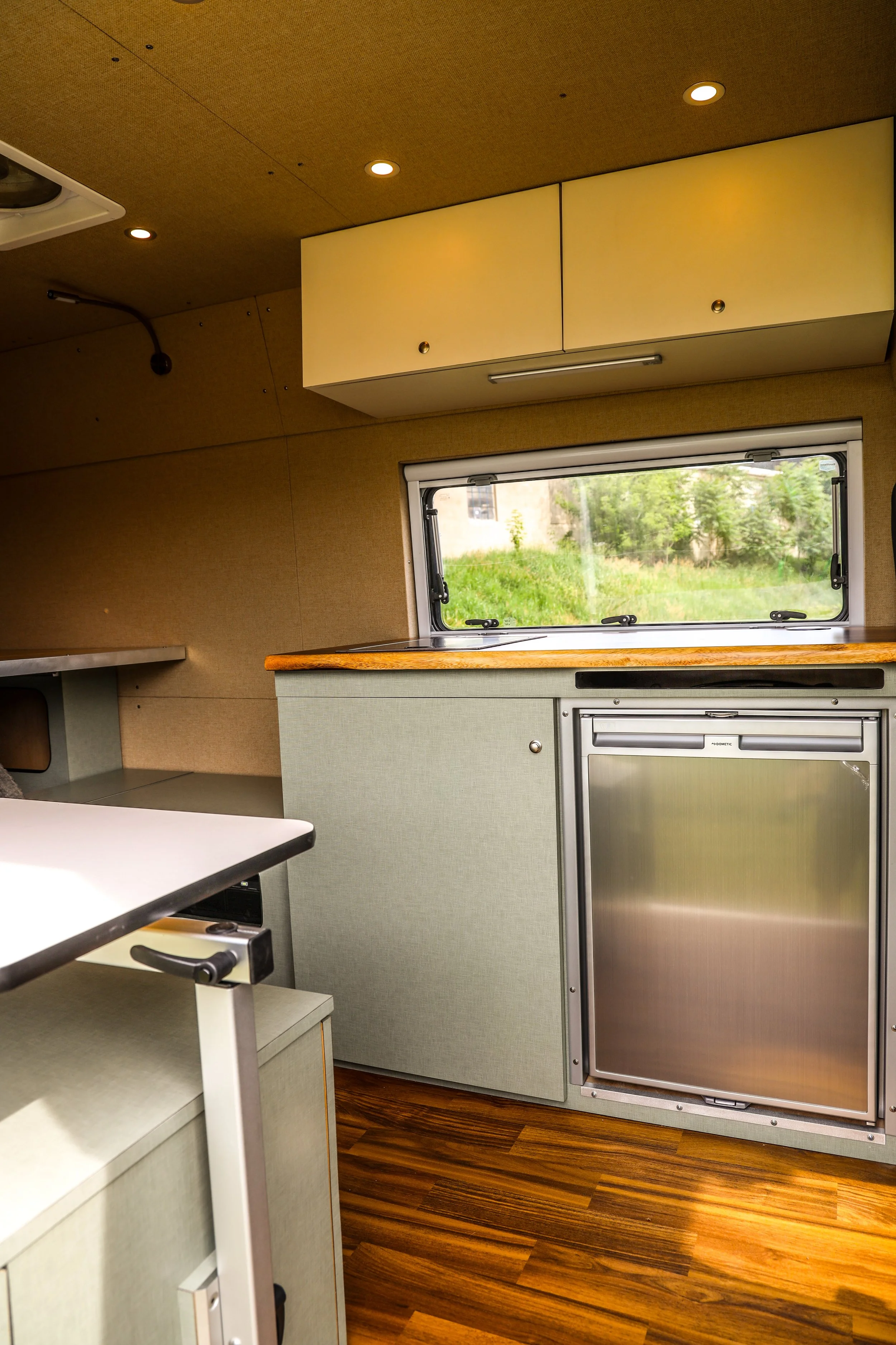 Interior of a camper van kitchen with a window, wooden countertop, beige cabinets, and stainless steel mini fridge, with wood-style flooring.