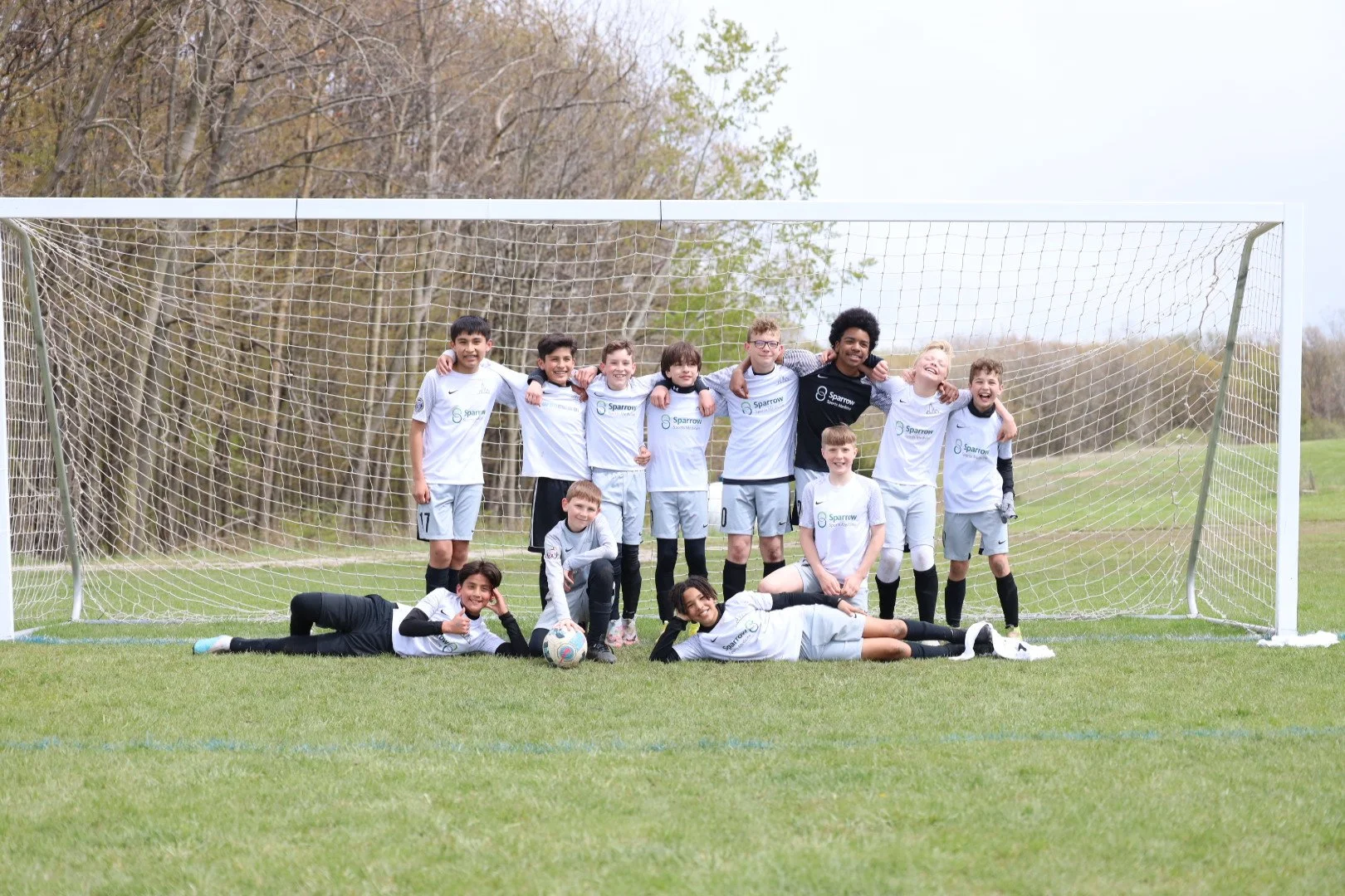 A group of young soccer players and a coach posing for a team photo on a soccer field, with some standing and some lying on the grass in front.