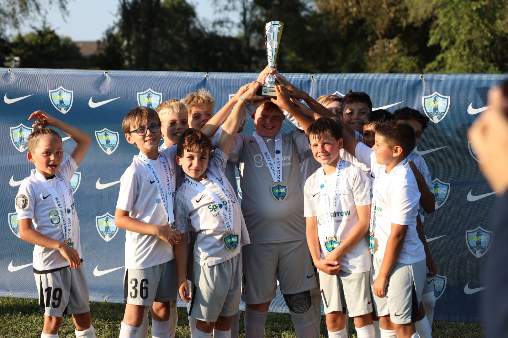 A group of young soccer players and their coach celebrating with a trophy, wearing white uniforms with medals around their necks, standing in front of a blue backdrop with team logos.