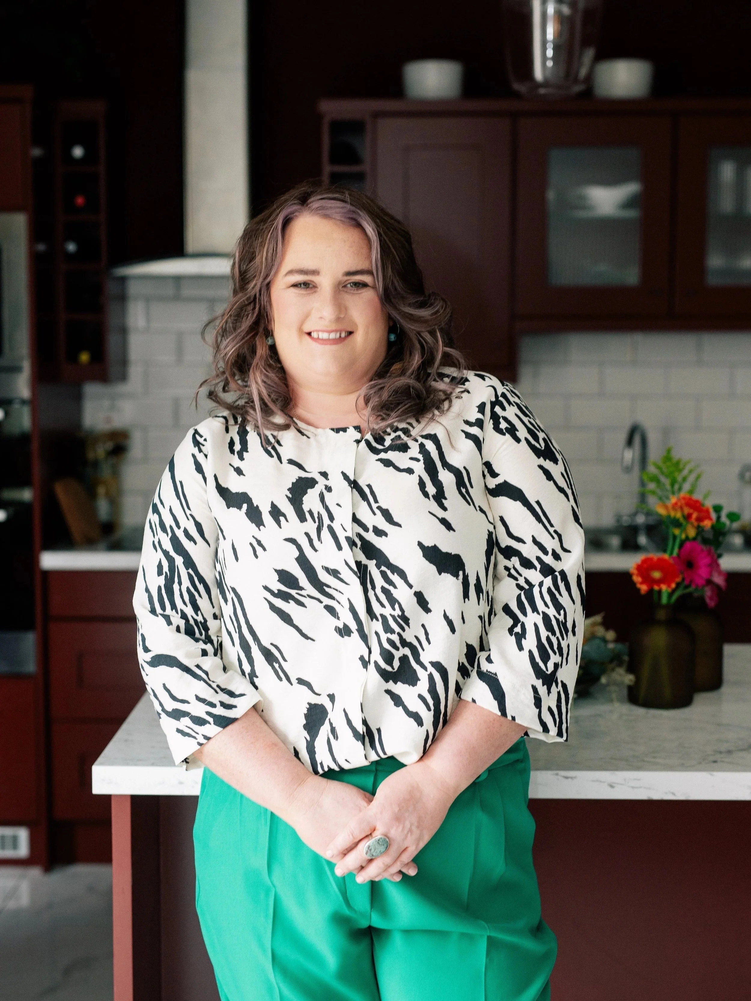 A woman with shoulder-length wavy hair, wearing a black-and-white animal print blouse and green pants, posing in a modern kitchen with dark wood cabinets and a white brick wall.