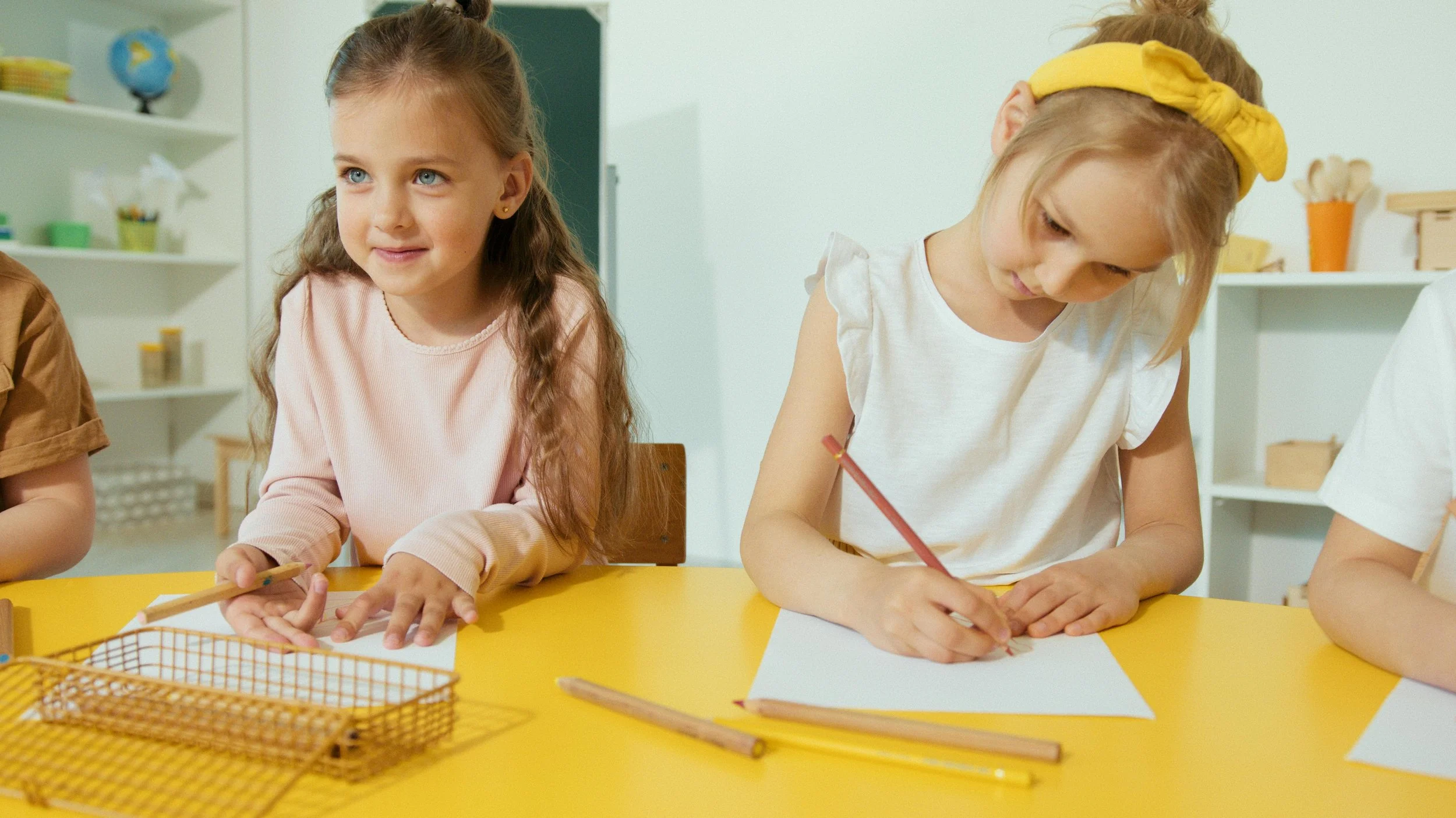 Two young girls sitting at a yellow table, one drawing with a red pencil on a white paper, the other looking to the side with a smile, in a classroom setting.