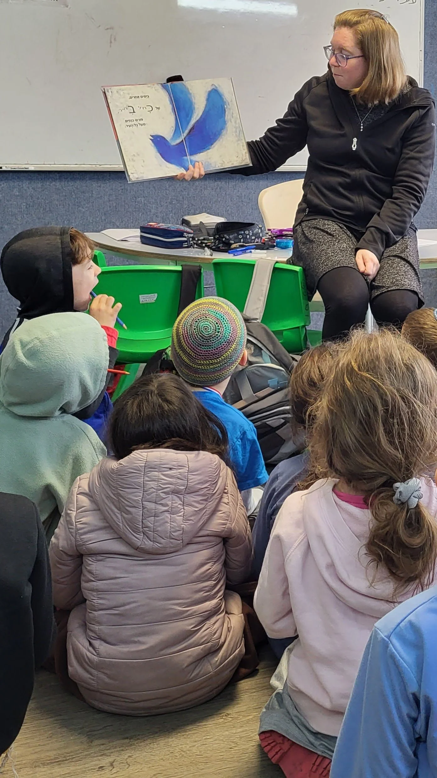 A woman in glasses and dark clothing is sitting on a stool, reading a colorful picture book to a group of children sitting on the floor in front of her. The children are wearing jackets and hats, and the setting appears to be a classroom or library.
