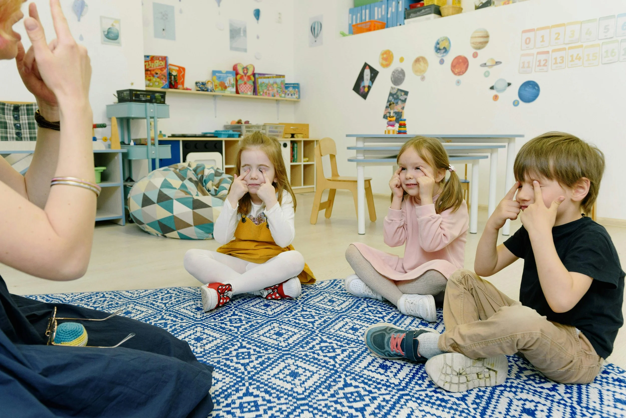 Three children sitting on a blue and white patterned rug, participating in a face exercise or eye exercise activity, guided by an adult.