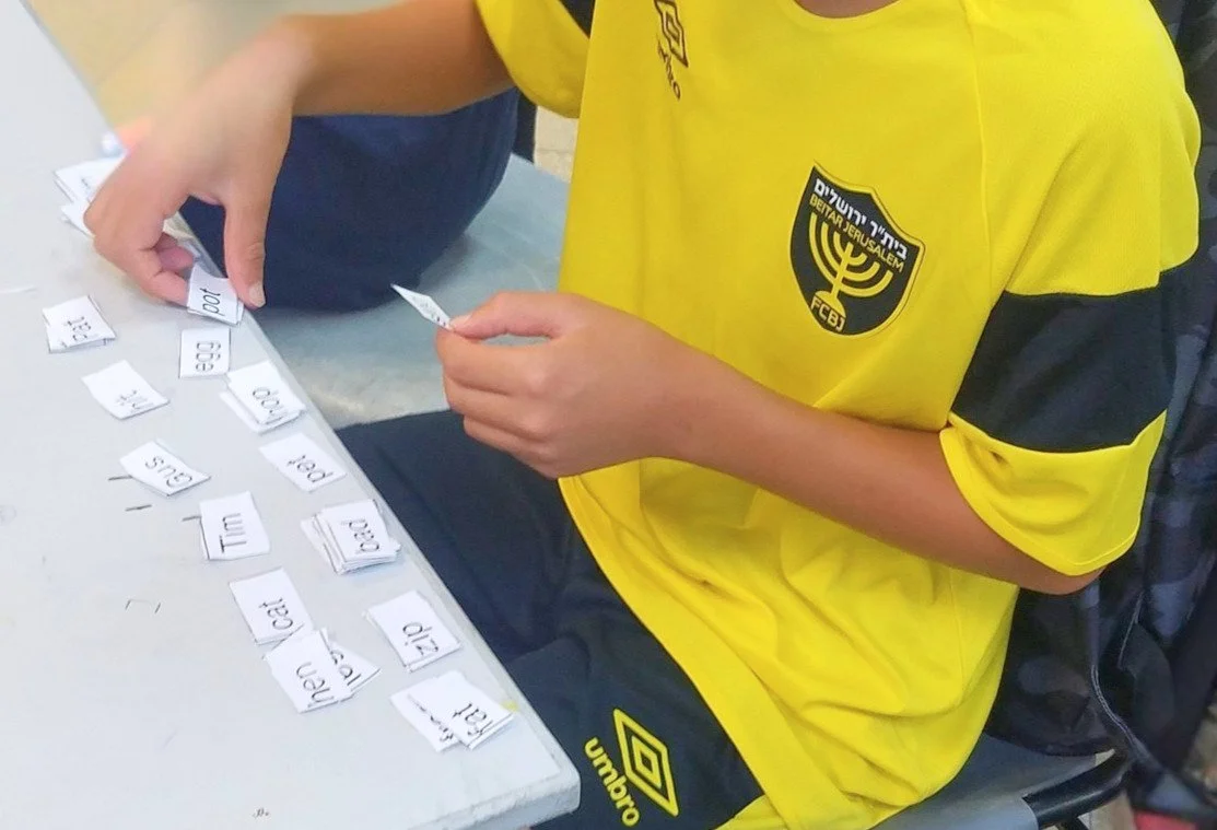 A person wearing a yellow FC Jerusalem sports jersey sitting at a table, using small pieces of paper with words to create sentences or phrases for a language activity.