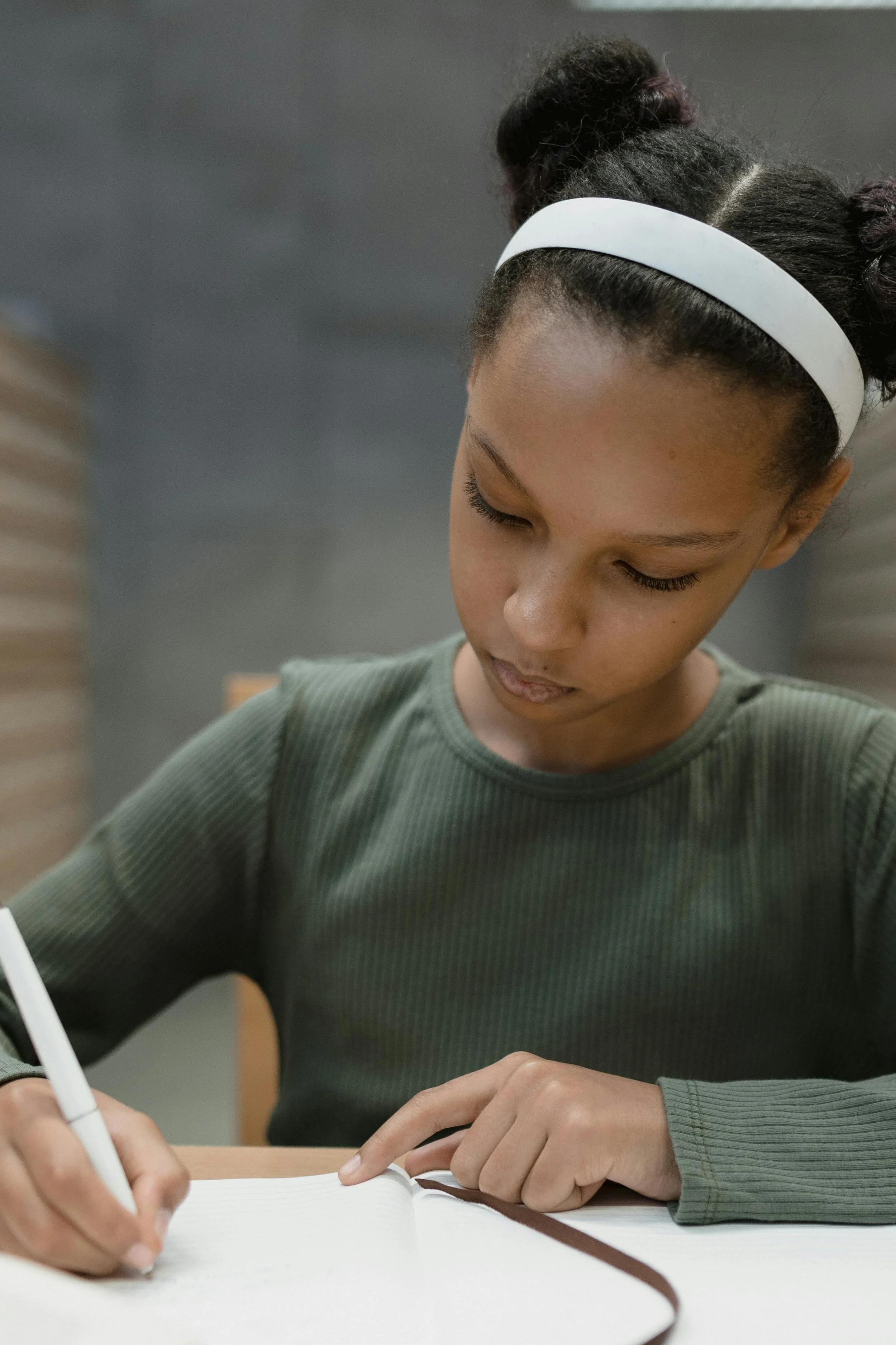 A young girl with braided hair secured by a white headband, writing in a notebook with a white pen while sitting at a wooden table.