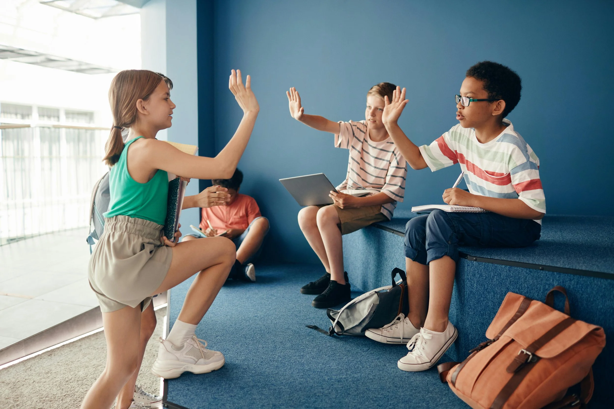 A female teacher giving a high five to a girl while two boys sit on a blue cushioned bench in a school hallway, one boy smiling with a notebook and the other on a tablet.