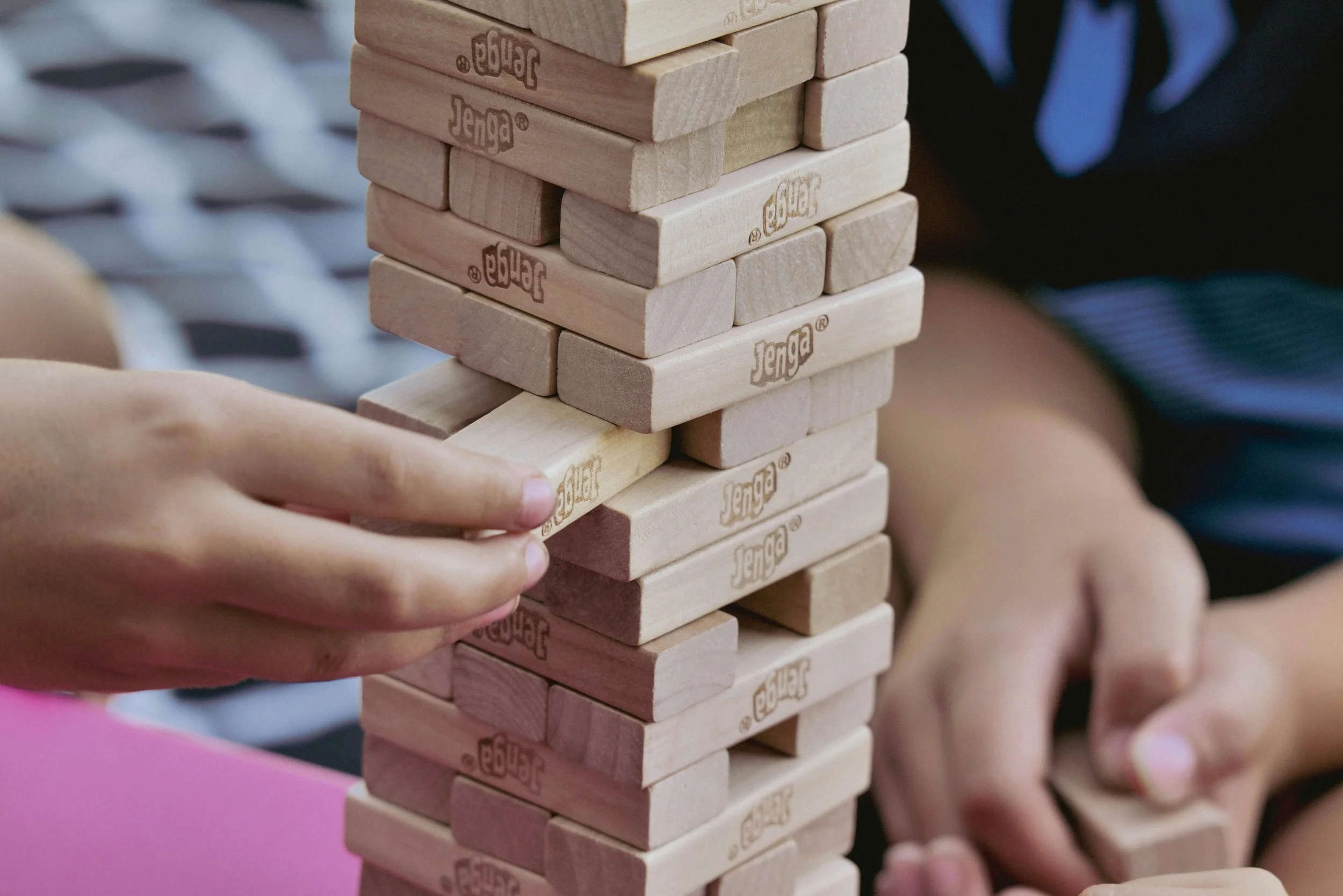 People playing Jenga with a tall tower of wooden blocks labeled Jenga, focusing on one person's hand carefully removing a block from the tower.