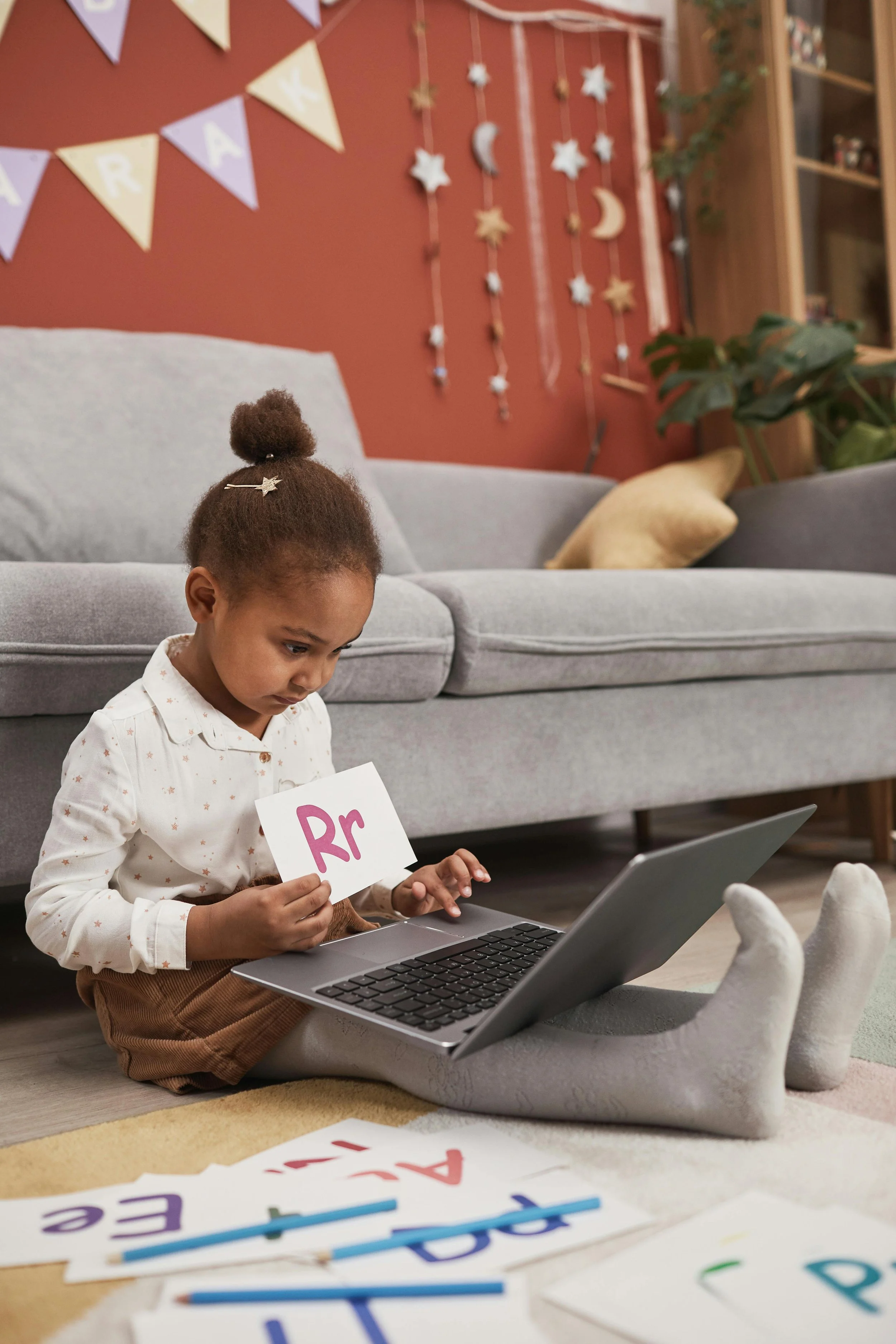 A young girl sitting on the floor with gray socks, holding up a card with 'Rr' written on it, in front of a laptop. Colorful alphabet flashcards are scattered on the floor nearby. Behind her is a gray sofa, decorated with pillows, and a red wall with hanging star and moon decorations.