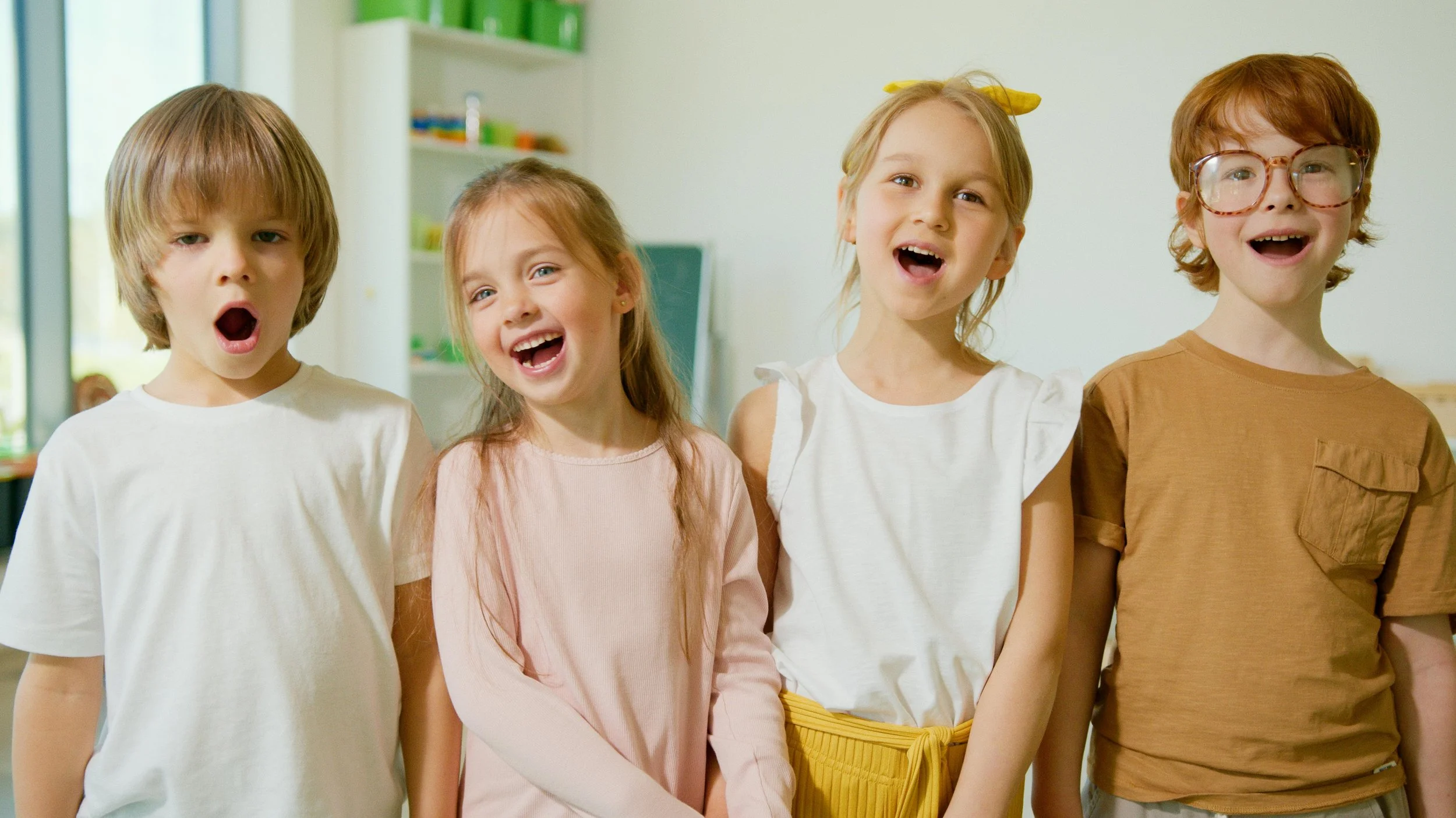 Four young children standing indoors, smiling and laughing. The children are dressed casually, with one girl wearing a yellow headband and another girl wearing large glasses. The background shows a bright room with shelves and a window.