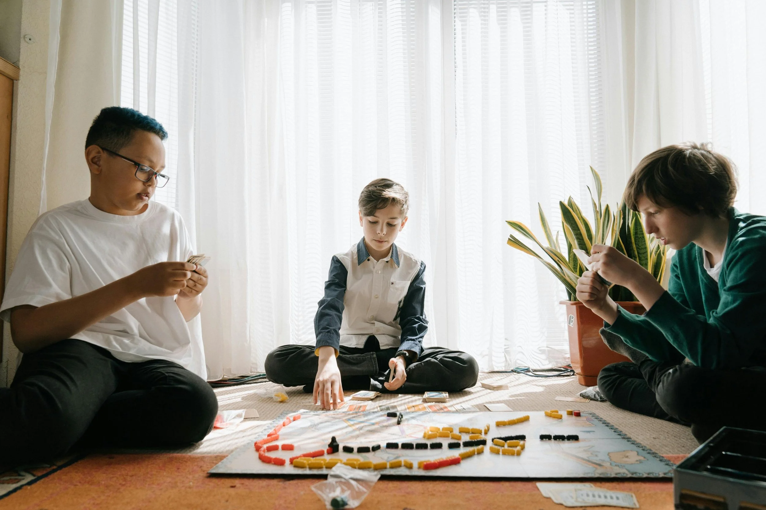 Three boys playing a board game on the floor in front of sheer curtains with sunlight coming through.