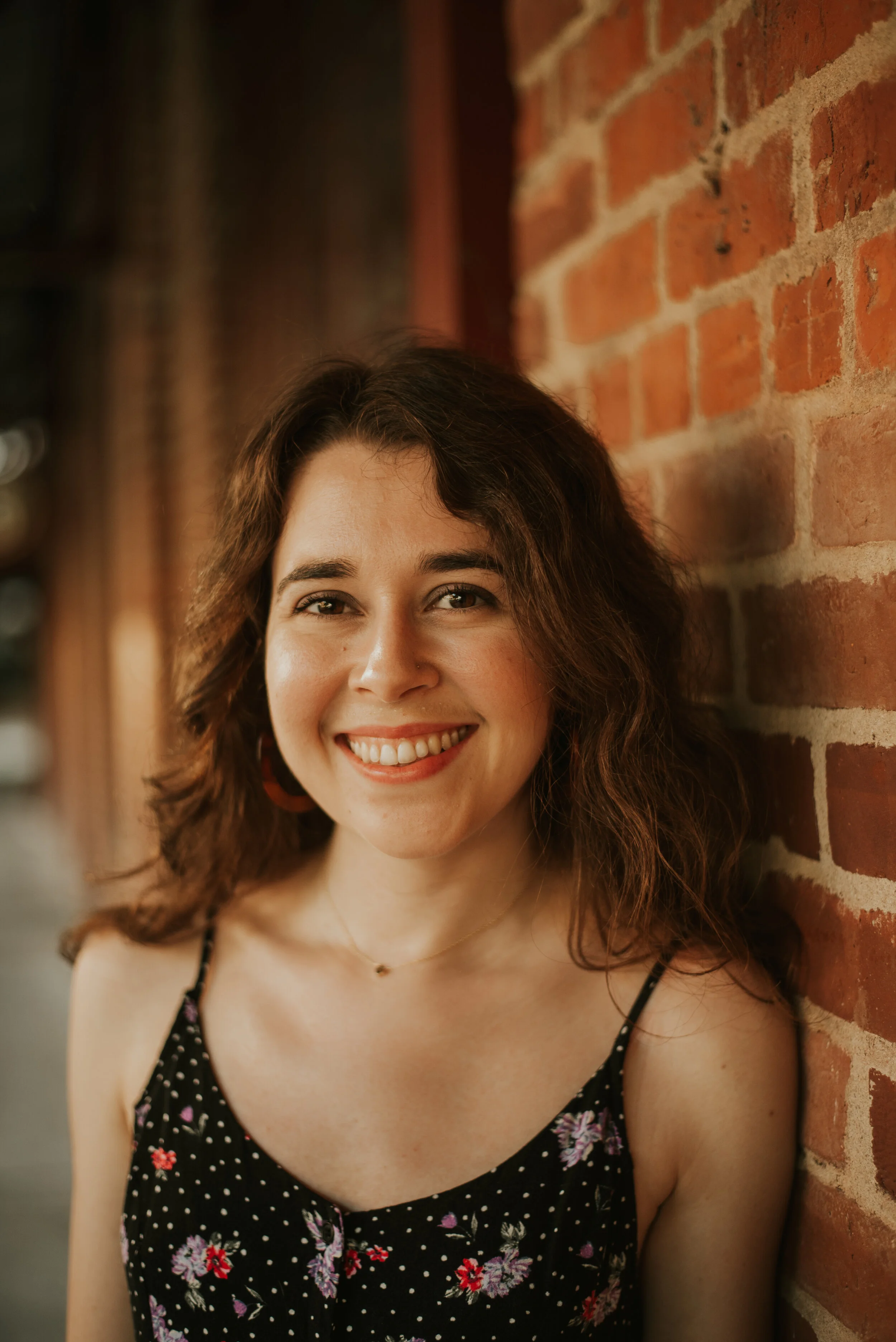 A young woman with brown, curly hair and a bright smile, standing against a red brick wall.