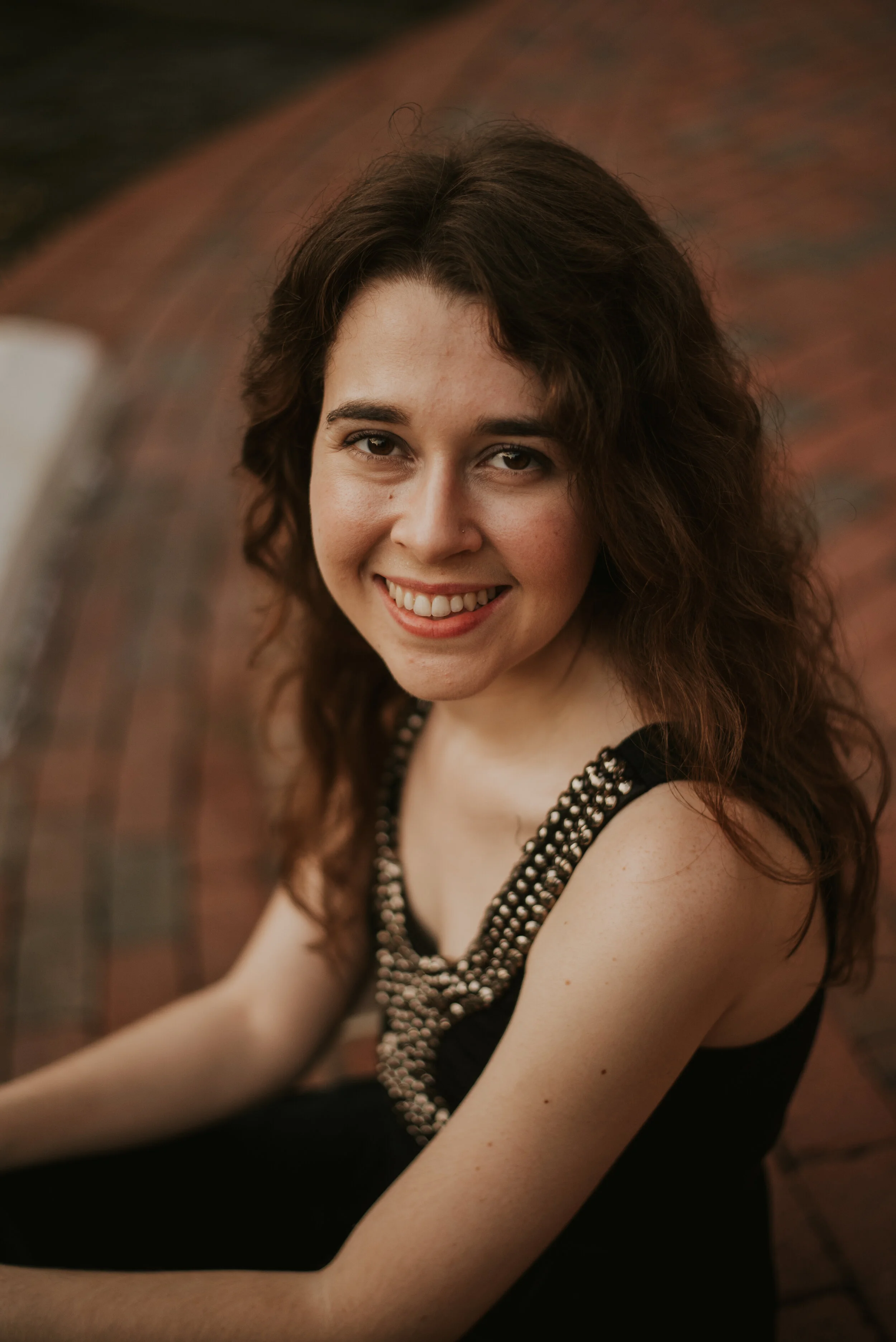 Close-up of a young woman with curly brown hair, smiling, wearing a black dress with pearl embellishments around the neckline, sitting in front of a brick wall.