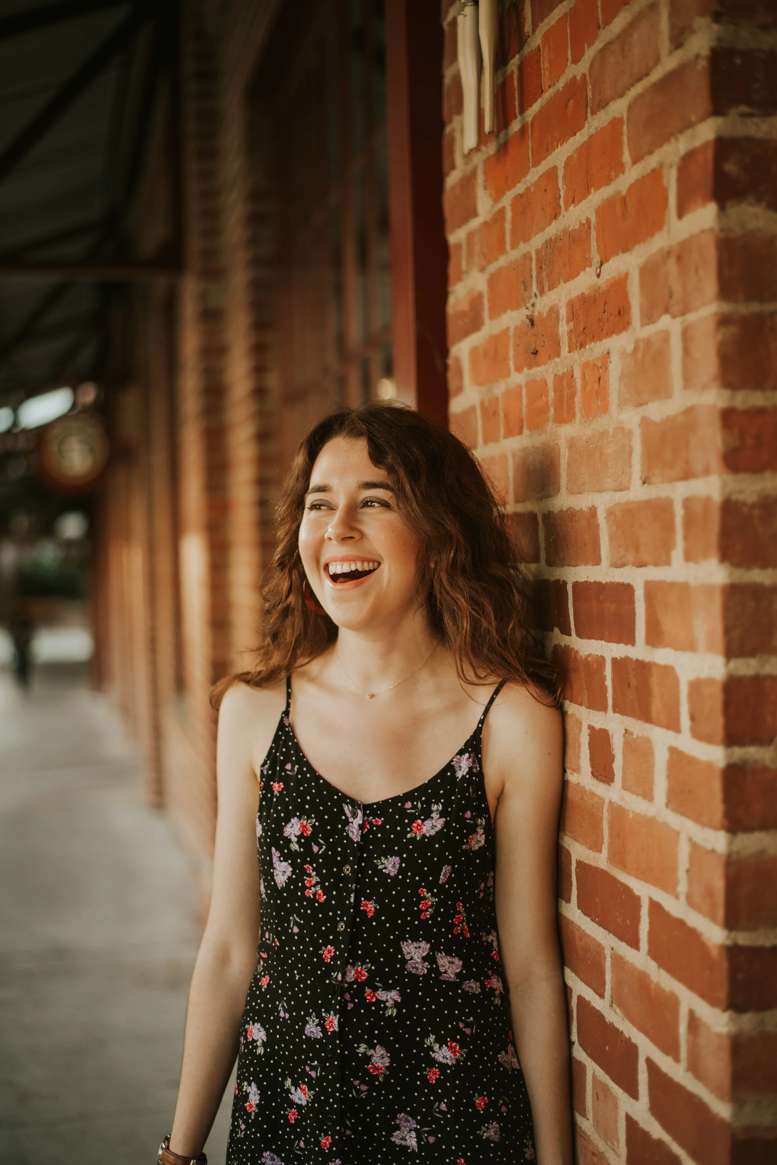 A young woman with dark brown wavy hair and light skin, wearing a black sleeveless dress with a floral pattern, is smiling and leaning against a red brick wall in an outdoor setting.