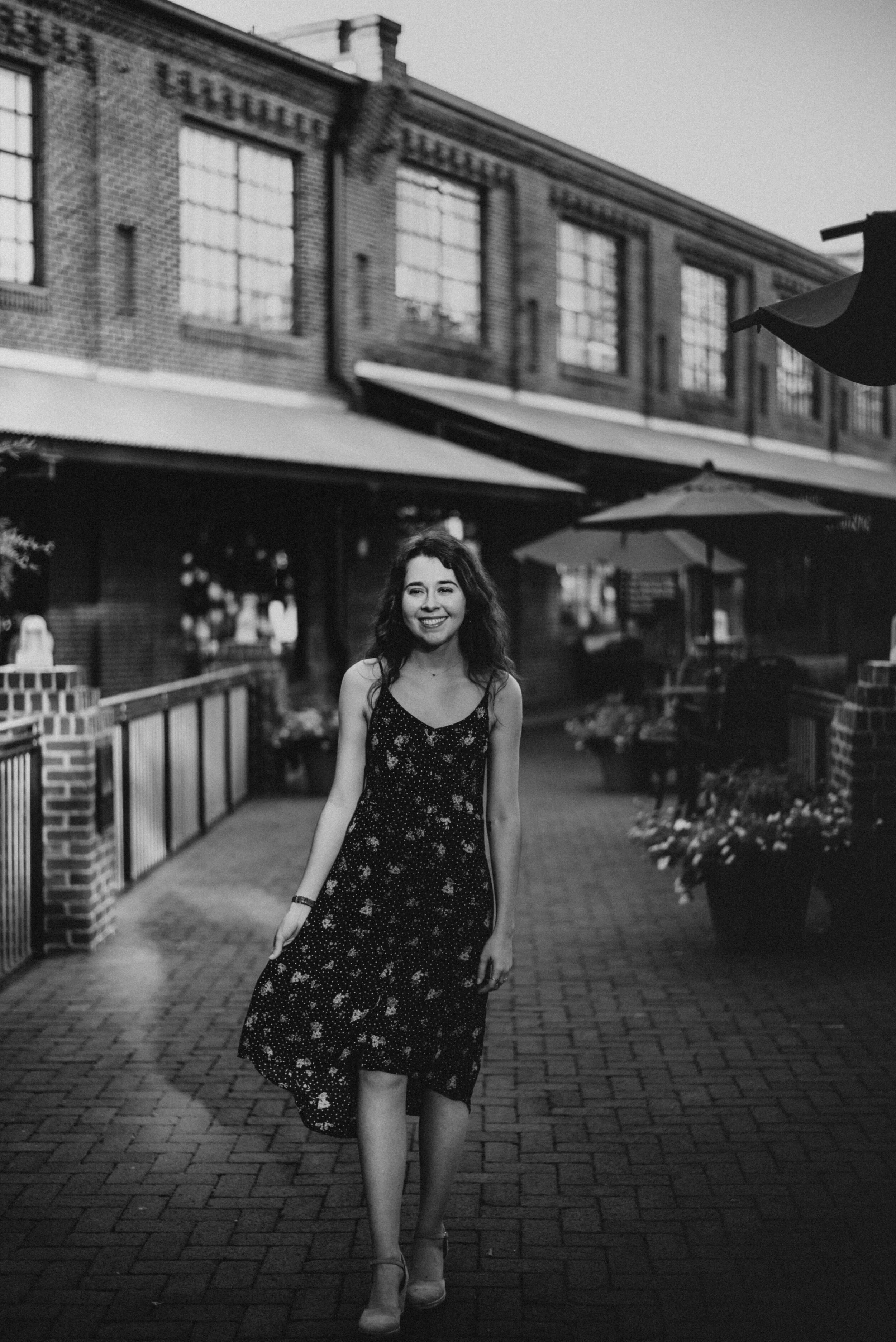A young woman wearing a sleeveless, floral dress, smiling and walking on a brick pathway in an outdoor urban setting during evening or night, with a brick building and umbrellas in the background.