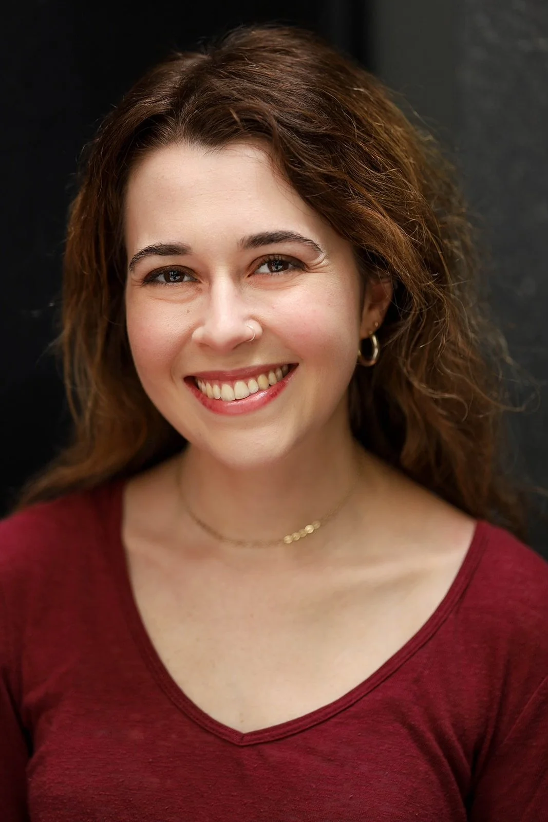 Close-up of a smiling woman with brown, wavy hair, wearing a red top, gold jewelry, and hoop earrings against a dark background.