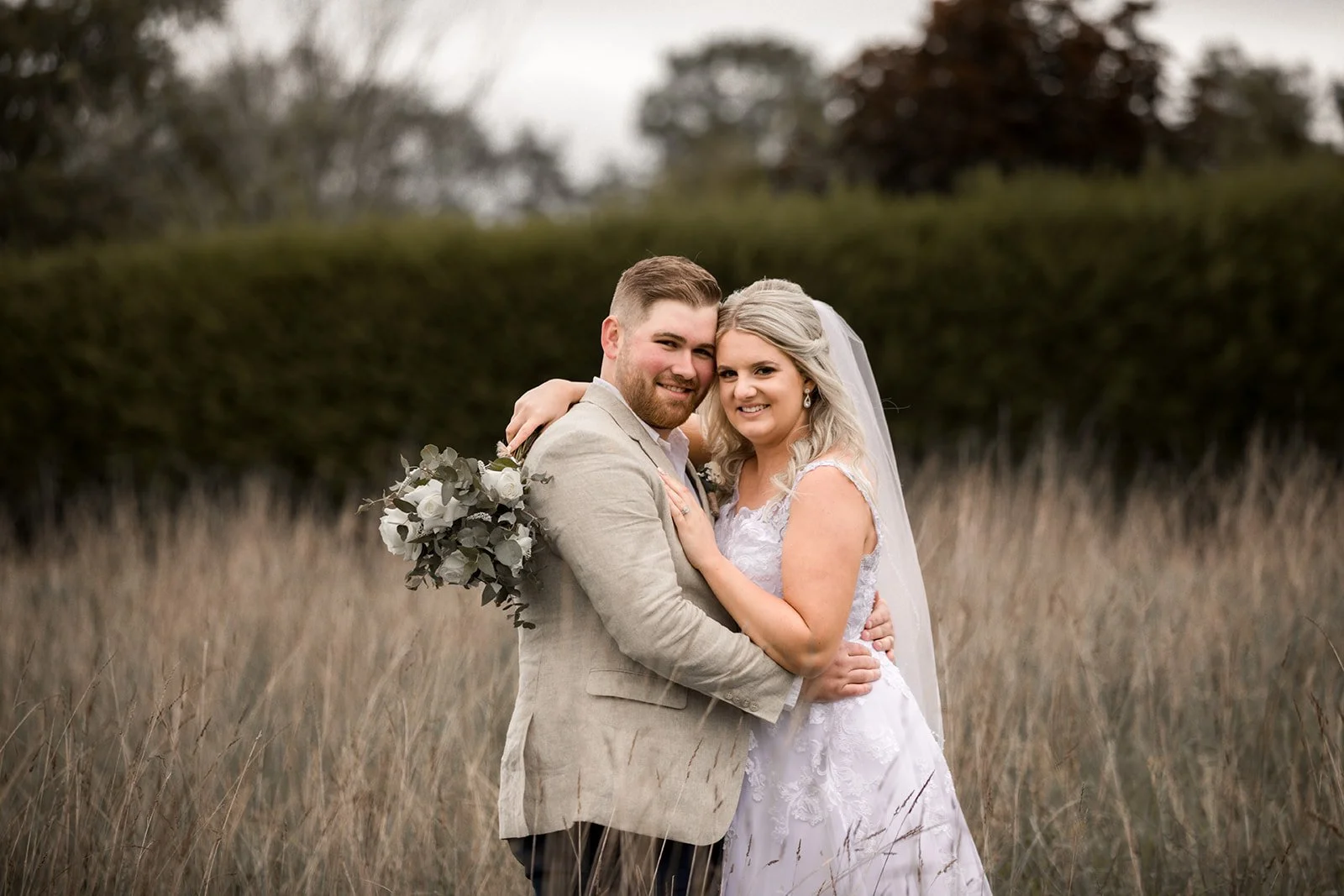 A newlywed couple standing in a field, embracing and smiling at the camera, with a blurred natural background and overcast sky.