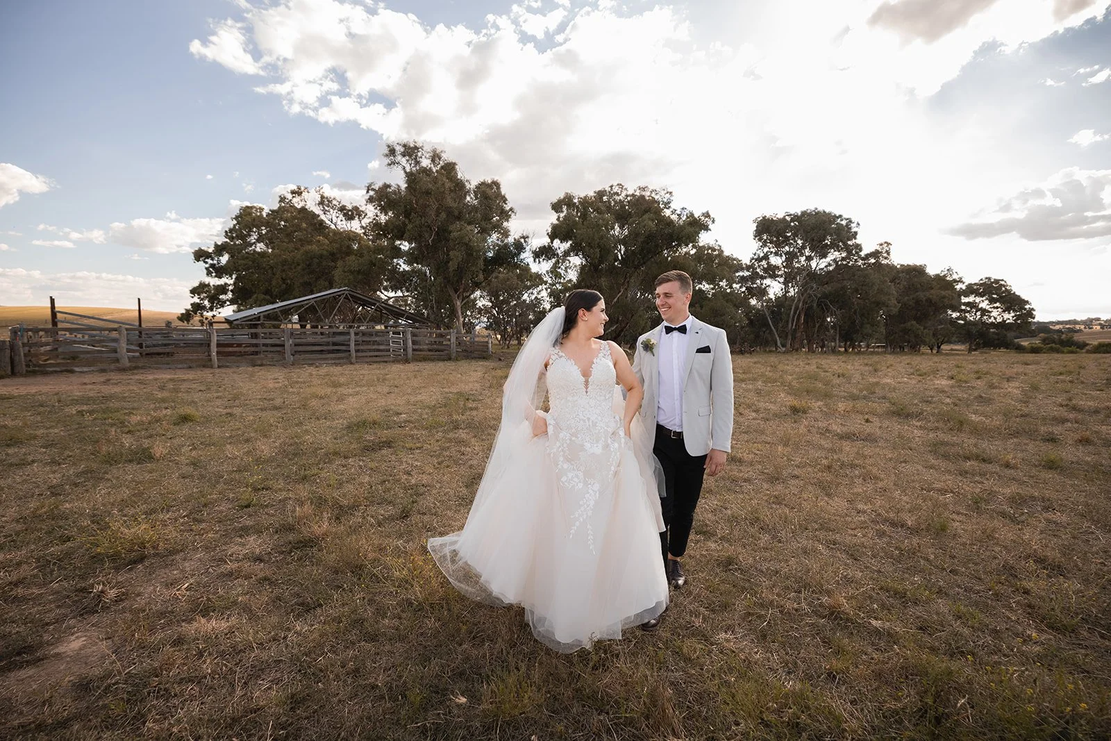 Bride and groom walking in open field at sunset, with trees and a wooden fence in the background.