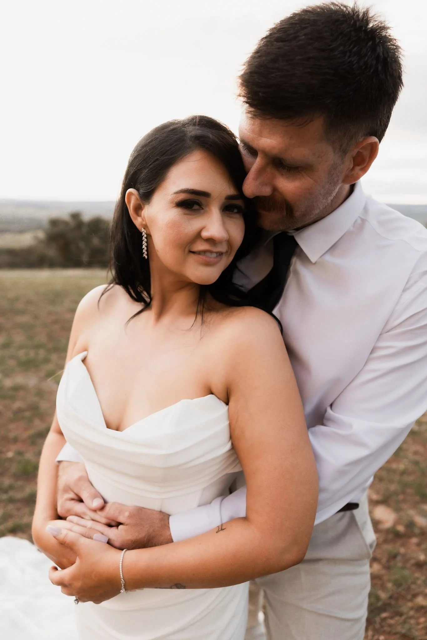A couple dressed in wedding attire embracing outdoors. The woman has dark hair, wearing earrings and a white off-shoulder wedding dress. The man has dark hair, beard, dressed in a white shirt with a black tie. They are holding each other closely, smiling softly, with a blurred landscape background.