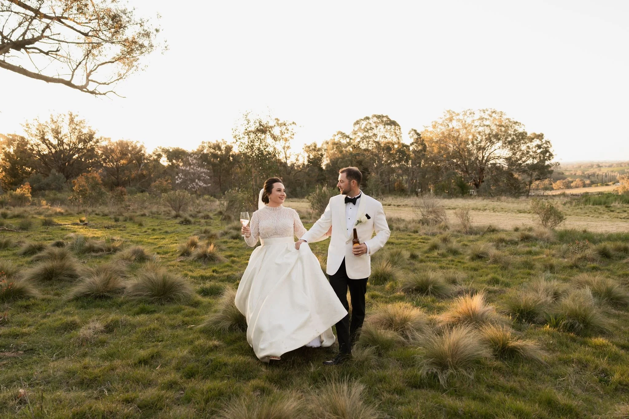 A bride and groom holding hands and walking through a grassy field with trees in the background during sunset.